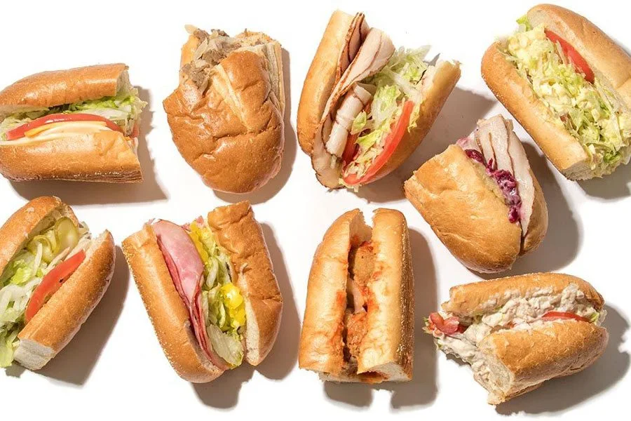 Top-down view of an assortment of Wawa hoagies on white background, with various fillings including turkey, ham, lettuce, tomato, and chicken salad. Emphasizes Wawa’s made-to-order deli sandwich options.