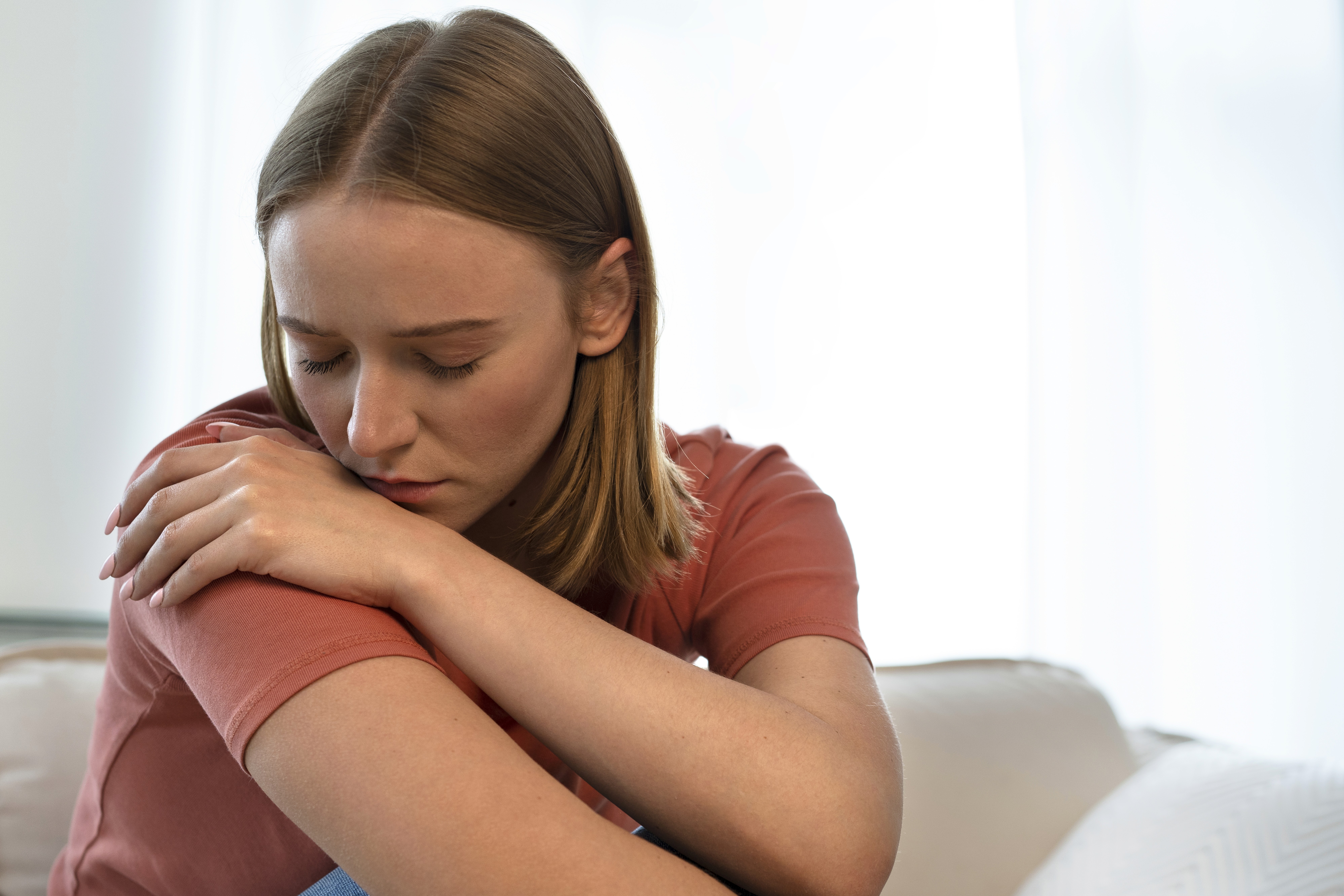 Close-up of a young woman looking down with a worried expression and arms crossed in a dimly lit setting.