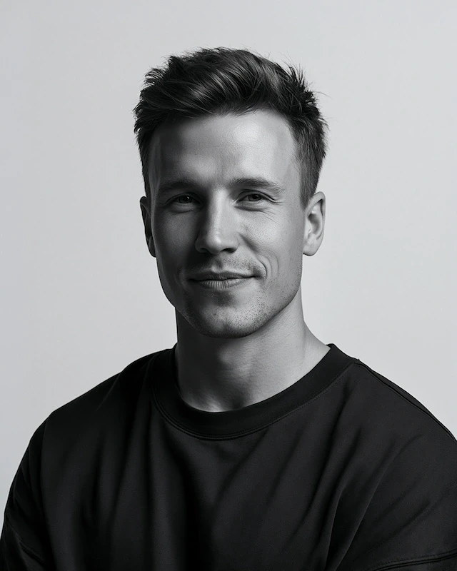 A black-and-white studio portrait of a confident man with short hair, wearing a dark crew-neck shirt against a gray backdrop.