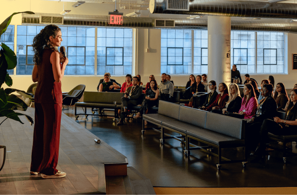 A speaker stands on a stage addressing an audience seated in a bright, modern space with large windows.