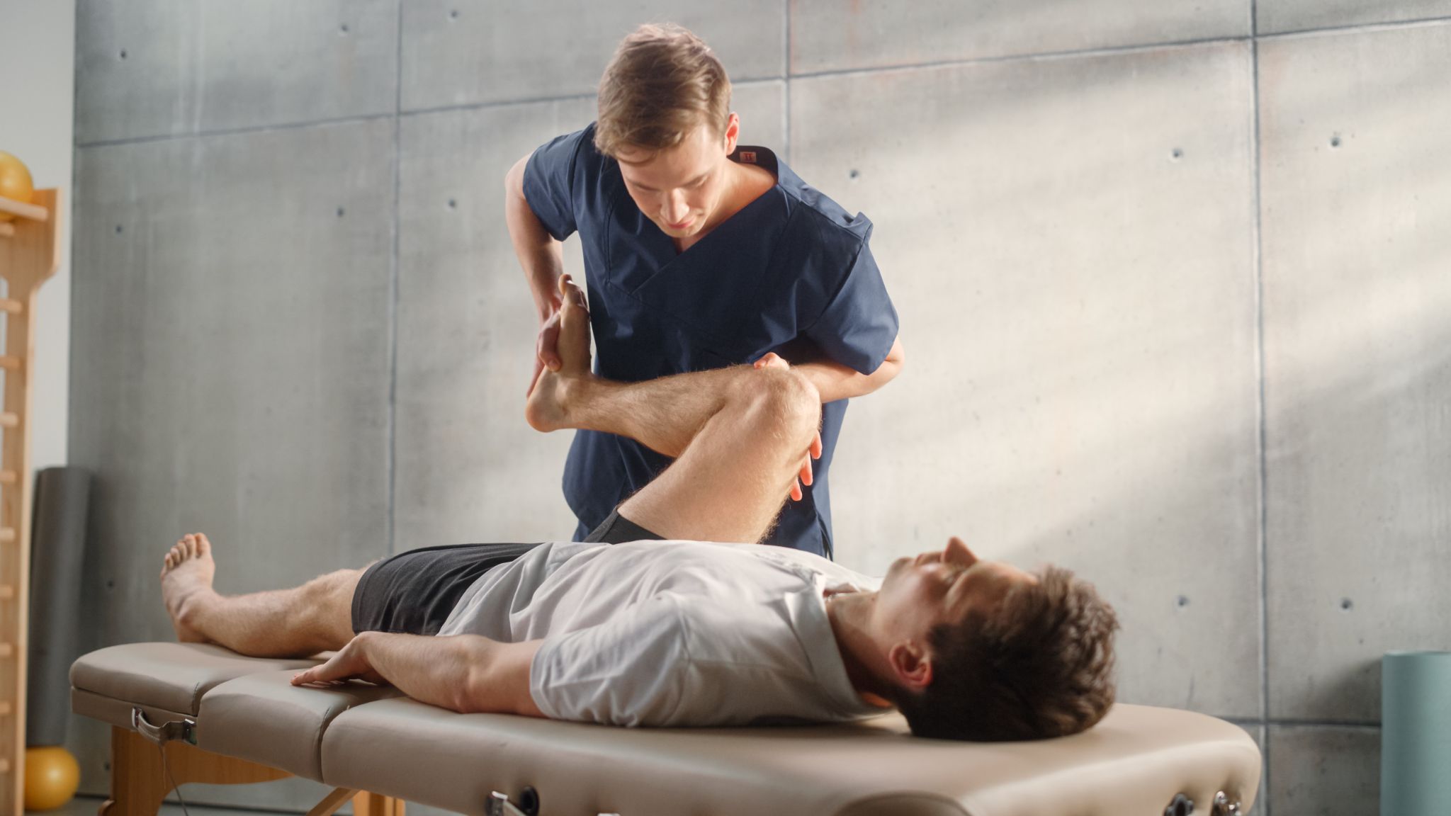 A physical therapist in blue scrubs performs a manual leg stretch and joint mobilization on a male patient lying on a treatment table.