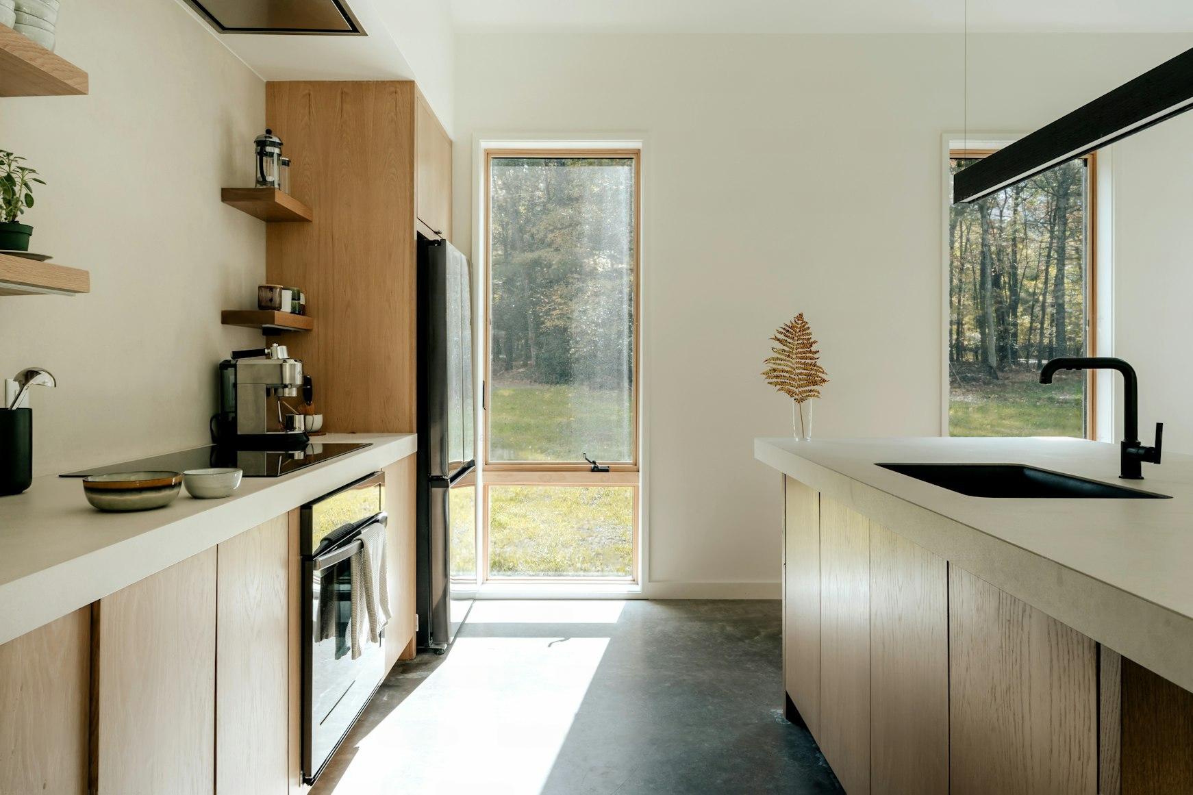 Compact off-grid kitchen with timber cabinetry, black matte tap, espresso machine, and large windows overlooking an autumn forest