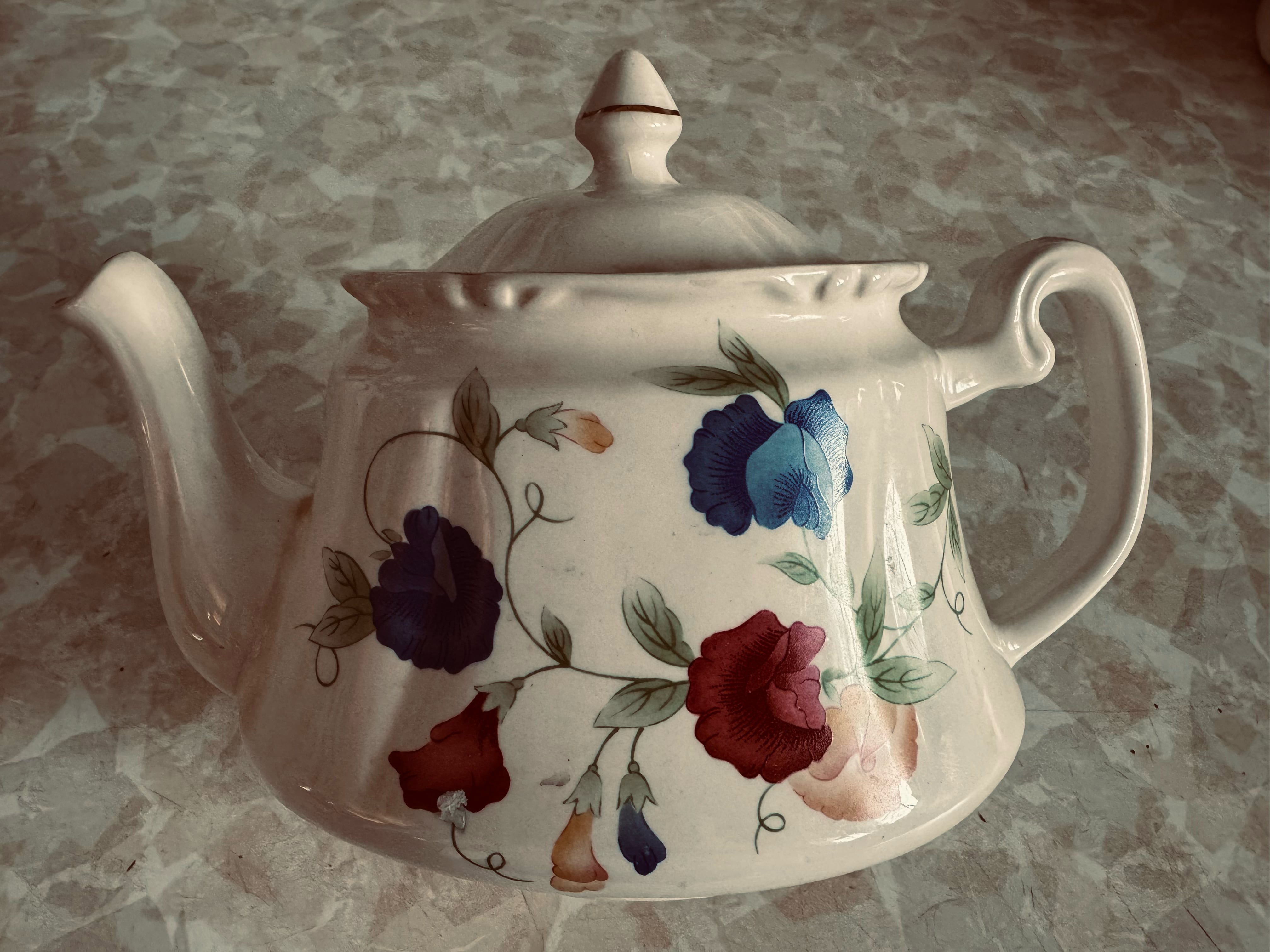 A cream coloured ceramic teapot with a painted design of sweetpeas against a mottled countertop