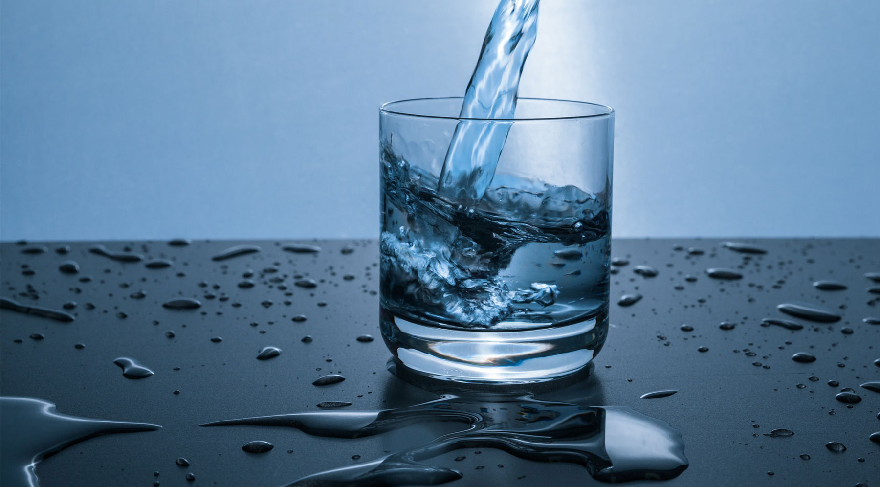 A clear glass filled with water sits on a surface, with droplets and light reflecting off it.