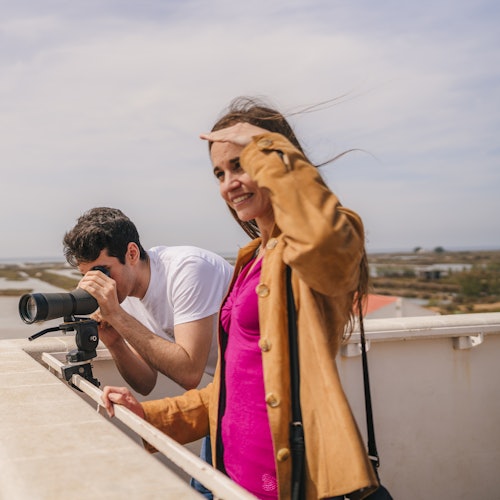 Ein Mann und eine Frau auf einem Dach; der Mann benutzt eine Kamera mit einem langen Objektiv, während die Frau ihre Augen mit der Hand abschirmt und lächelt.