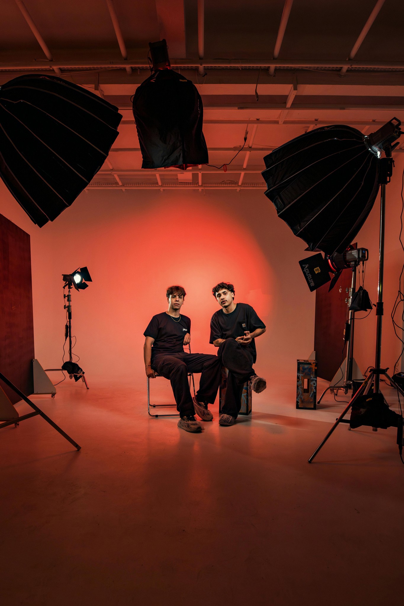 Two men pose in a photo studio surrounded by lighting equipment under a dramatic red glow.