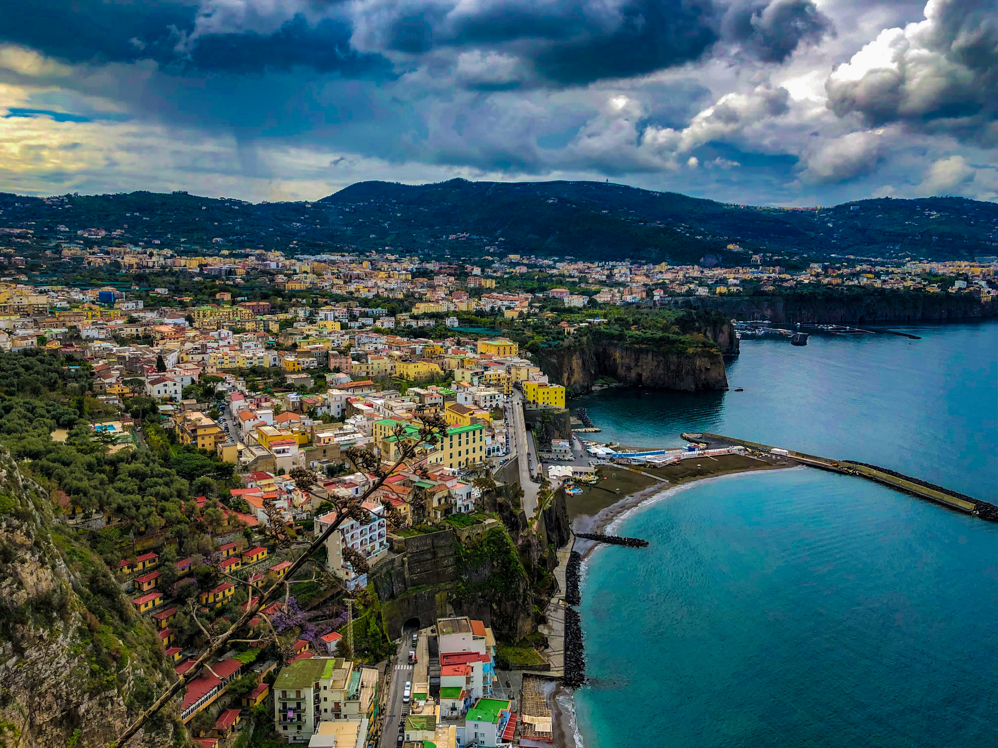 Una vista panoramica di Sorrento, Italia, che mostra edifici colorati lungo la costa, scogliere drammatiche e una baia tranquilla con acque turchesi limpide sotto un cielo nuvoloso.