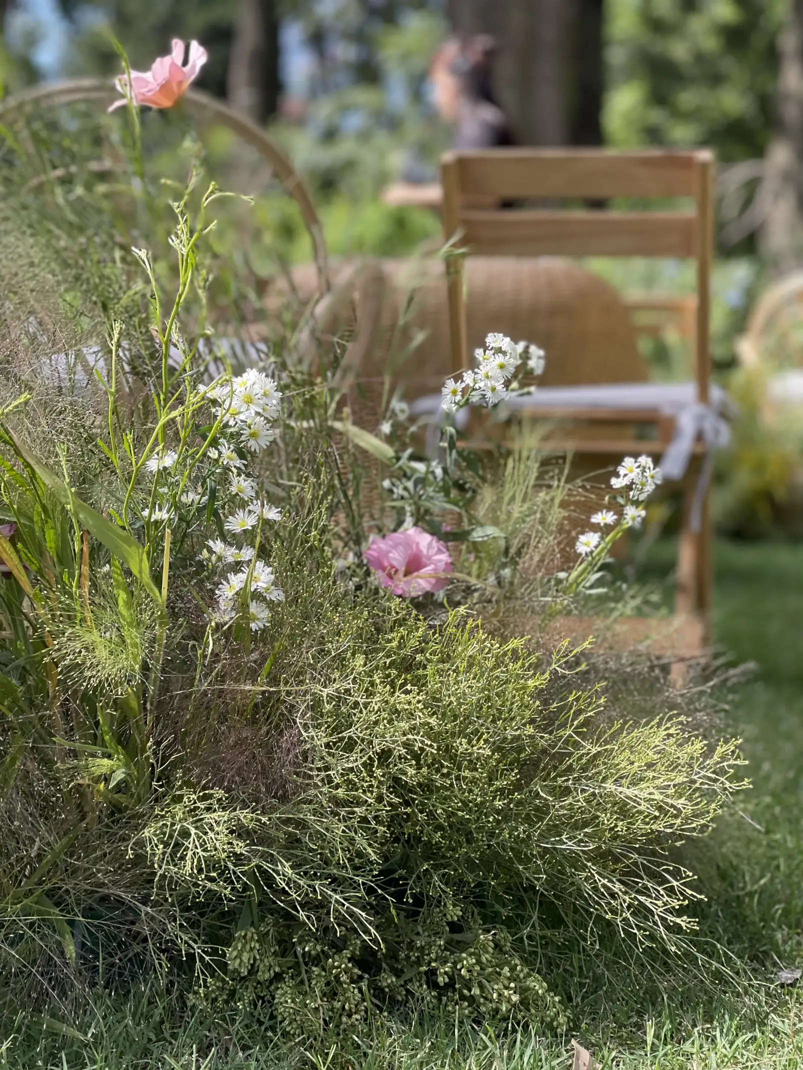 Arreglo floral silvestre con flores rosadas y blancas junto a sillas de madera para ceremonia natural al aire libre.