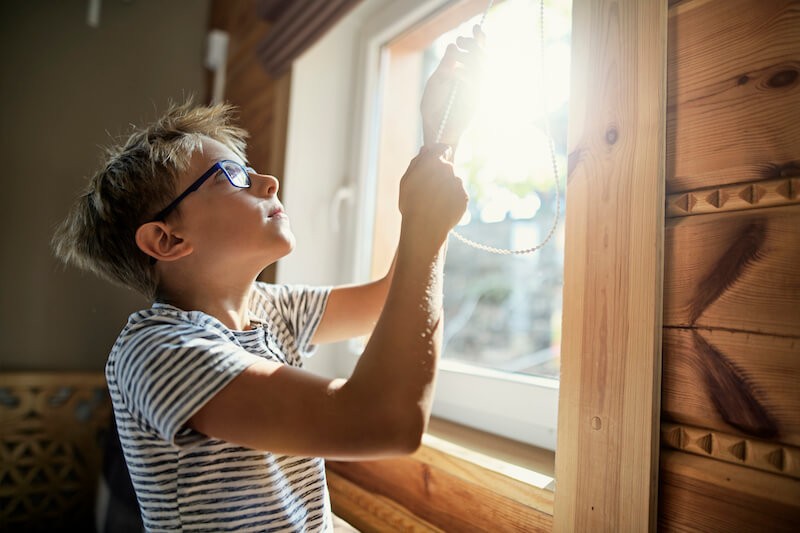 young boy opening faux wooden blinds to let sunlight in