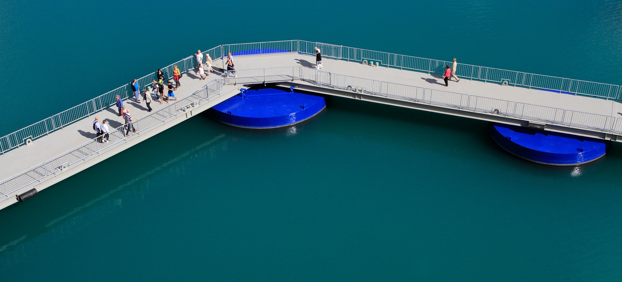 Pedestrians walking across the SeaWalk floating pier with blue pontoons beneath, demonstrating safe passenger access over calm harbor waters.