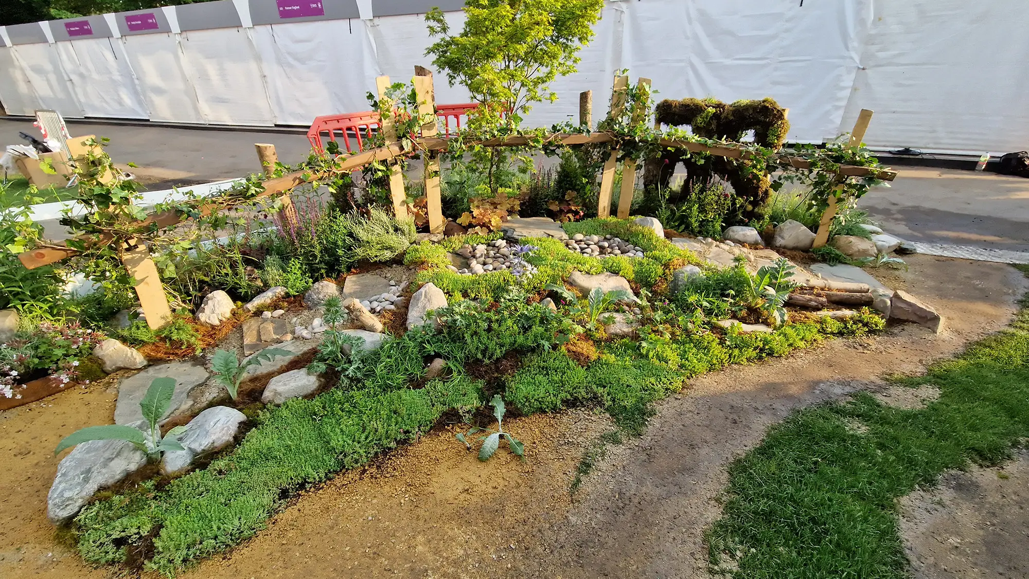 A scenic garden with various plants, stones, and pathways, set against a clear sky.
