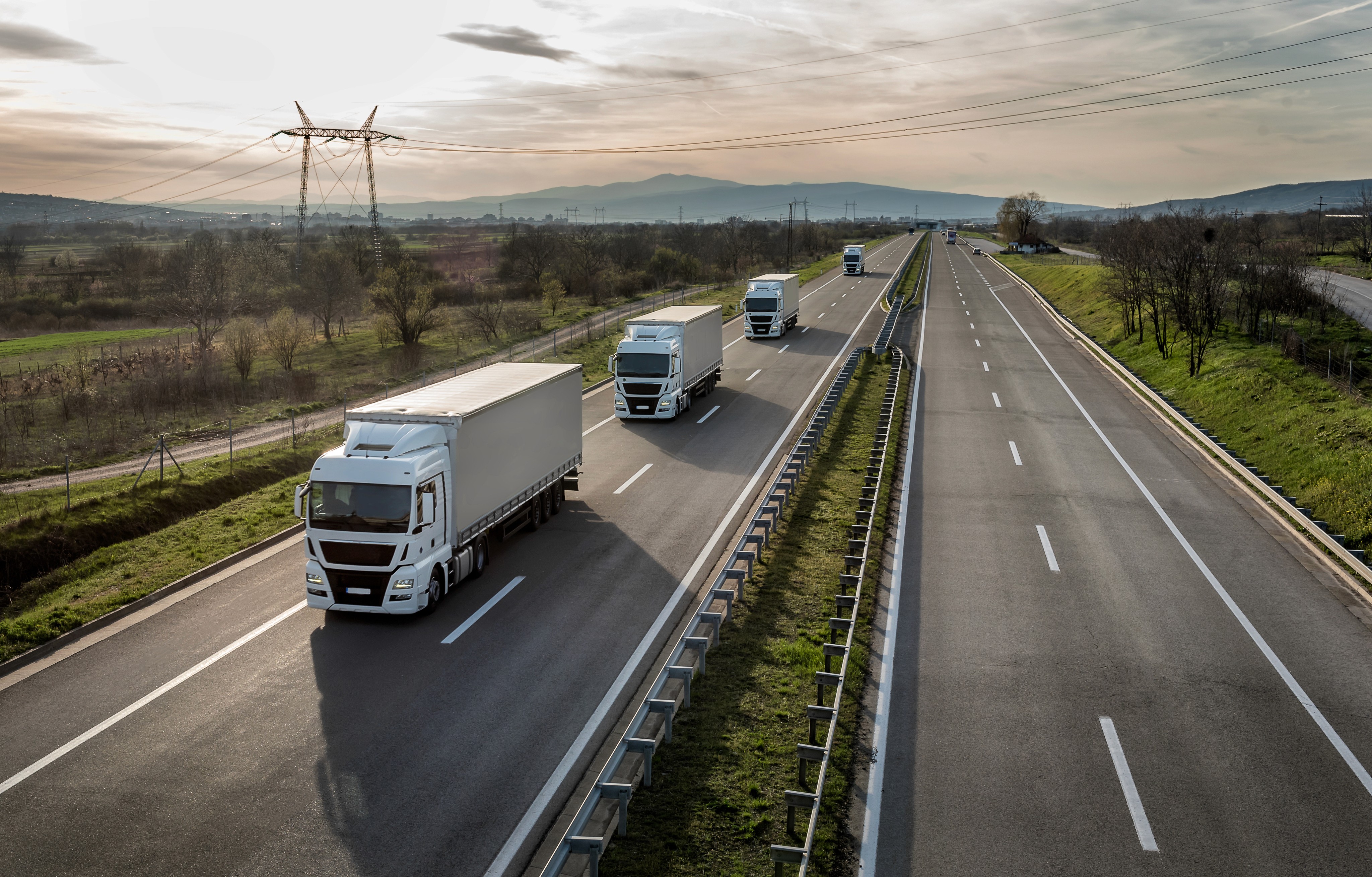 A line of trucks driving on a highway, set against a backdrop of fields and a cloudy sky.