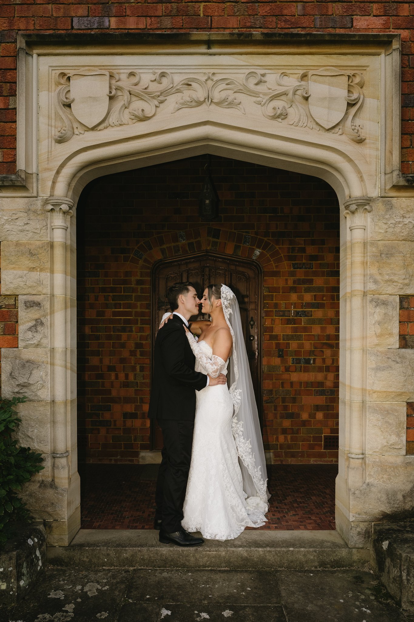 Bride and groom embrace in a carved sandstone archway of a historic brick building.