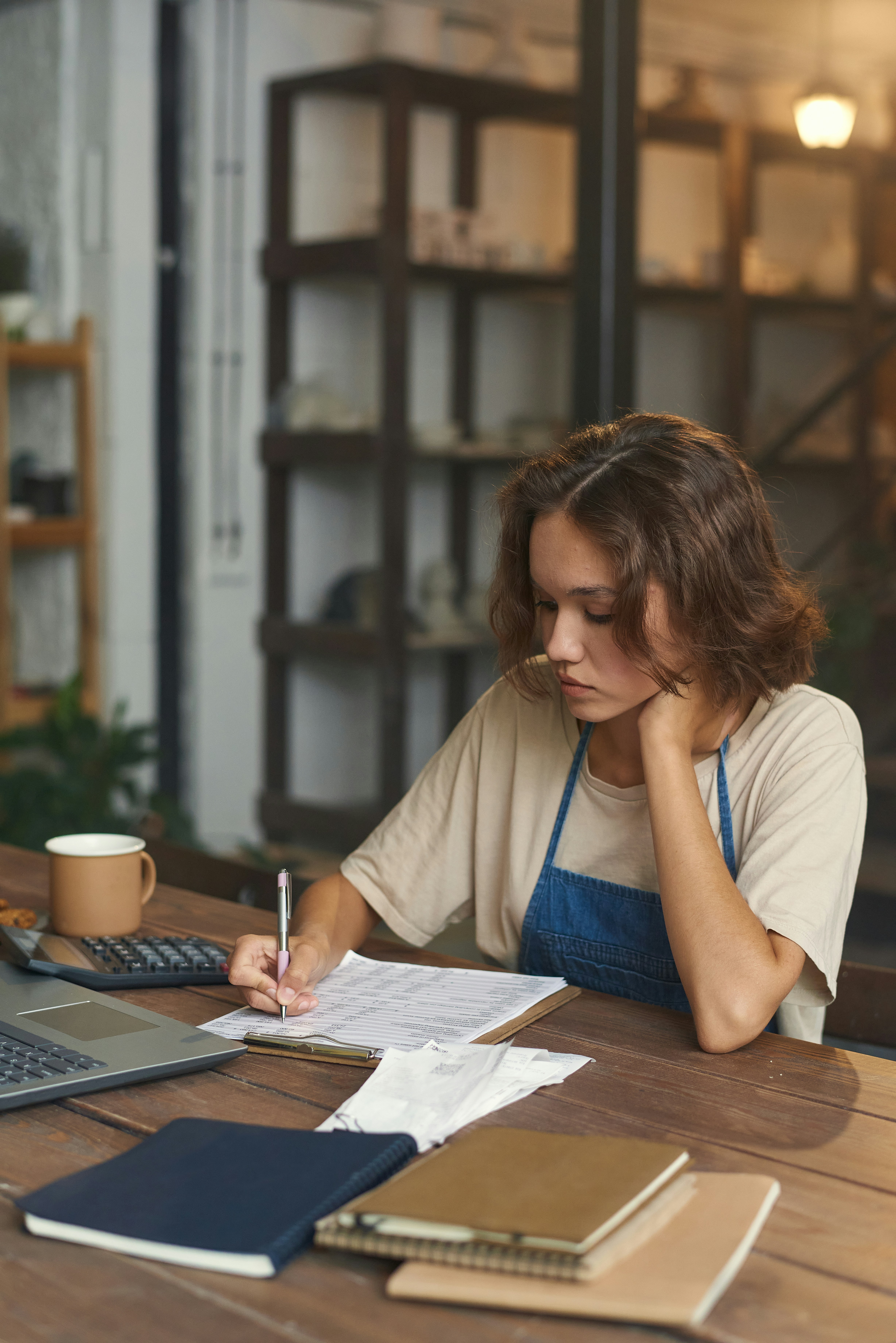 Small business owner in a denim apron sitting at a wooden desk reviewing paperwork, with a calculator, laptop, receipts, and notebooks in front of her, warm shop interior in the background.