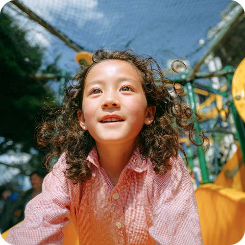 A young child with curly hair and a pink shirt smiles joyfully while playing on a sunny playground slide. The background shows vibrant playground equipment.