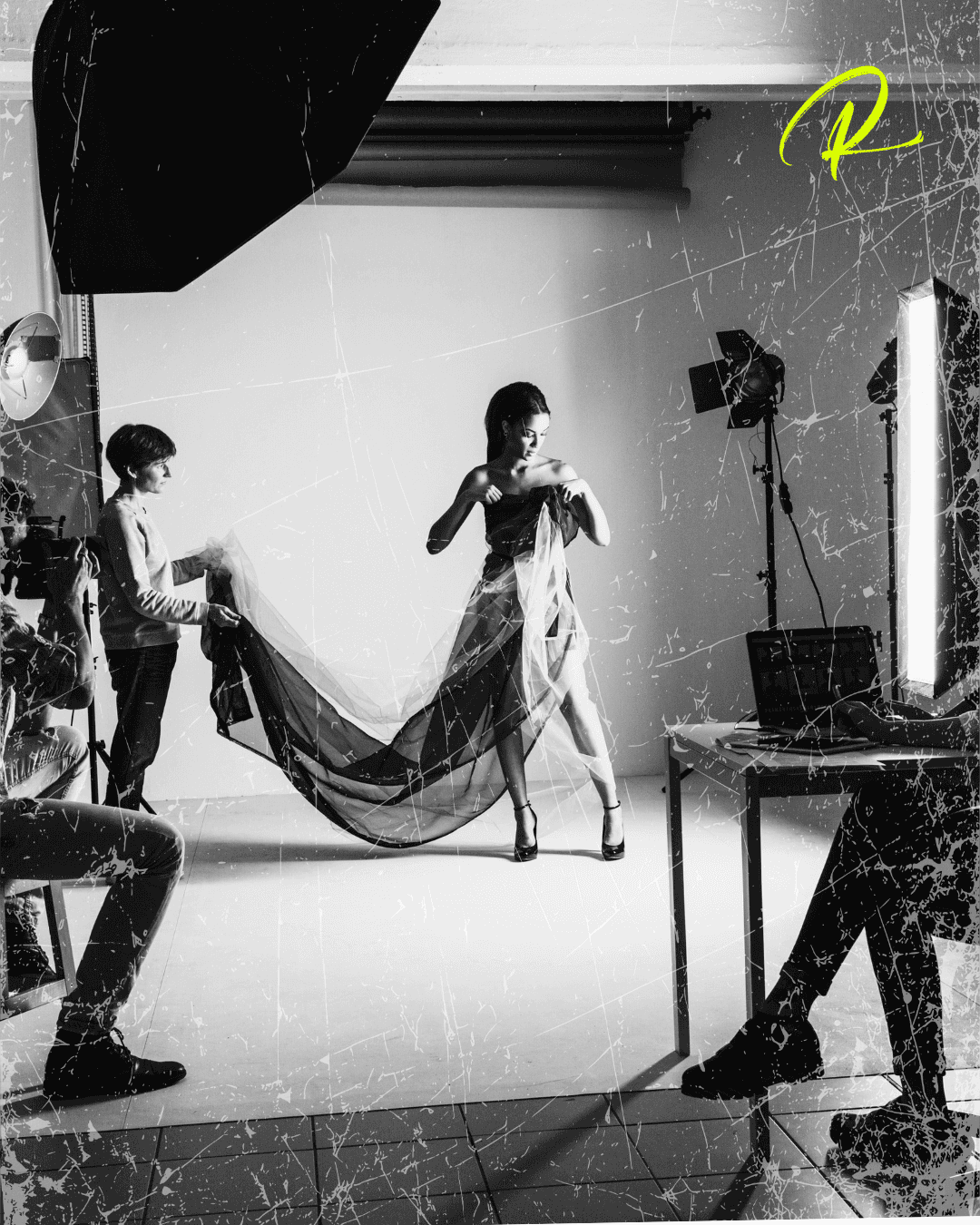 Black-and-white fashion shoot with a model posing in a studio as crew members adjust fabric.