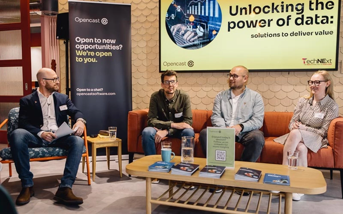 Four speakers sit on a stage sofa during a panel titled “Unlocking the power of data.” A low table with books and water sits in front, reinforcing a talk‑based format.