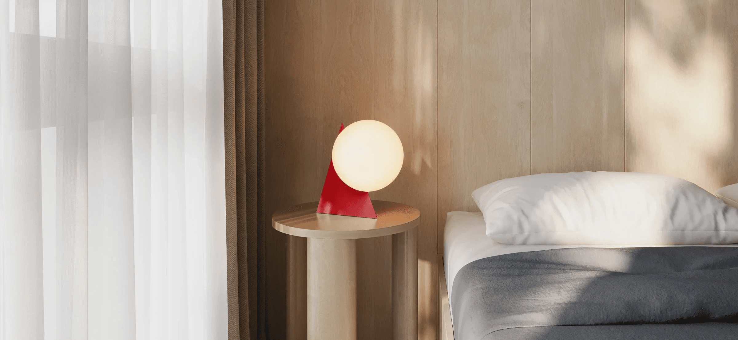 Red triangle table lamp with a glowing globe on a round wood side table beside a bed and sheer curtains