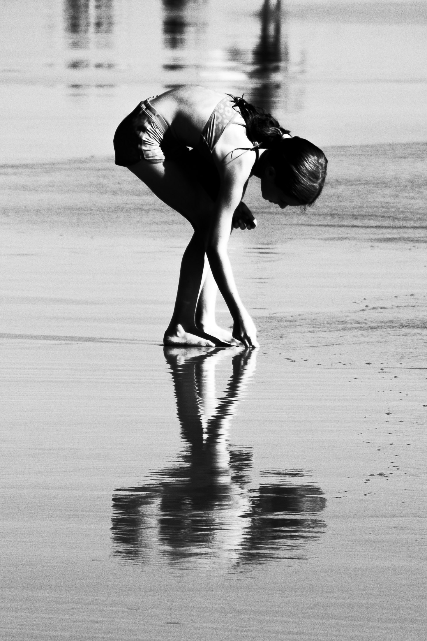 Girl bending down at the shoreline, reflected in wet sand, black and white photograph.