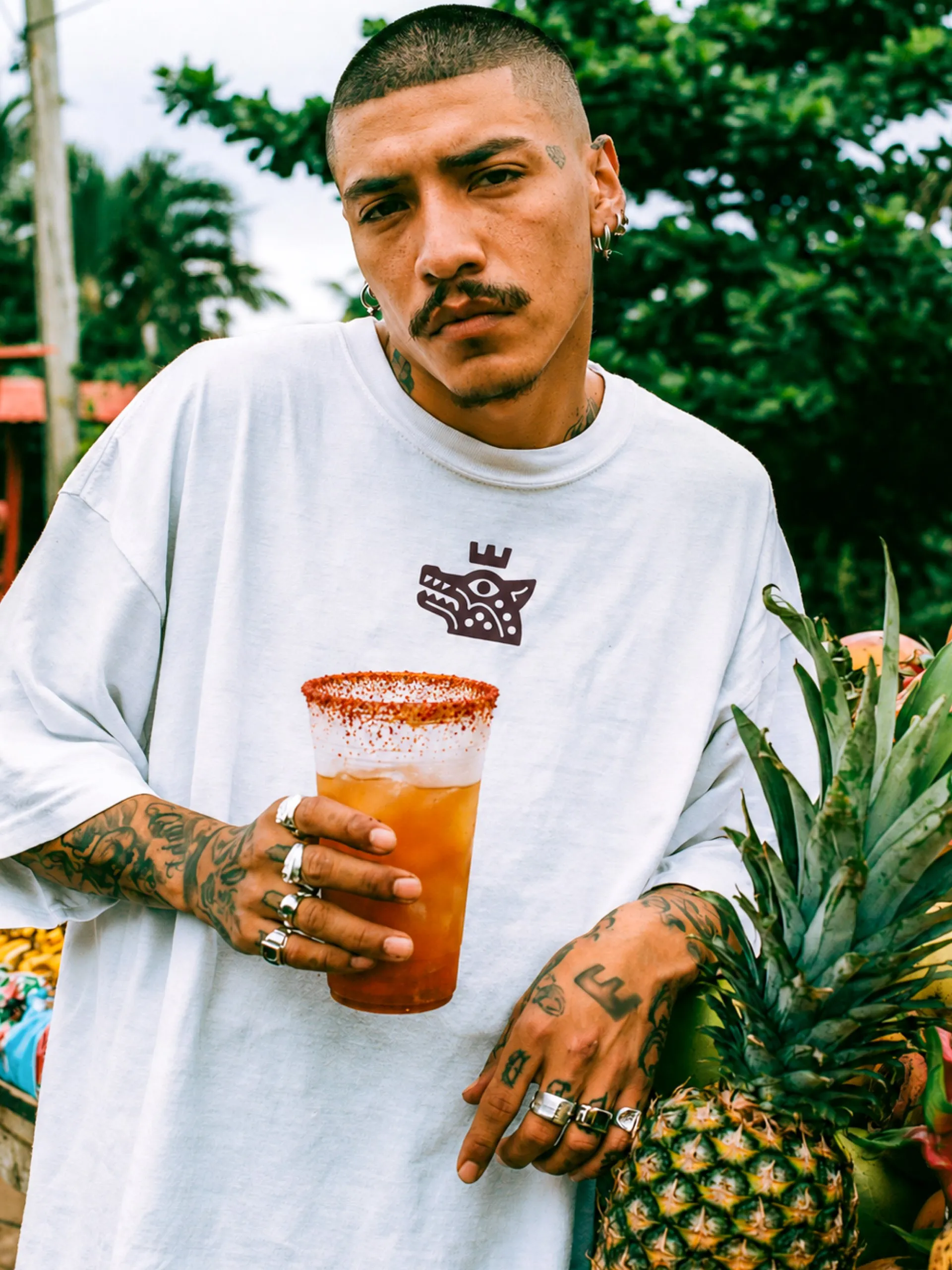 Tattooed man in a white Sagrada Michelada T-shirt holding a michelada beside a pineapple, brand apparel
