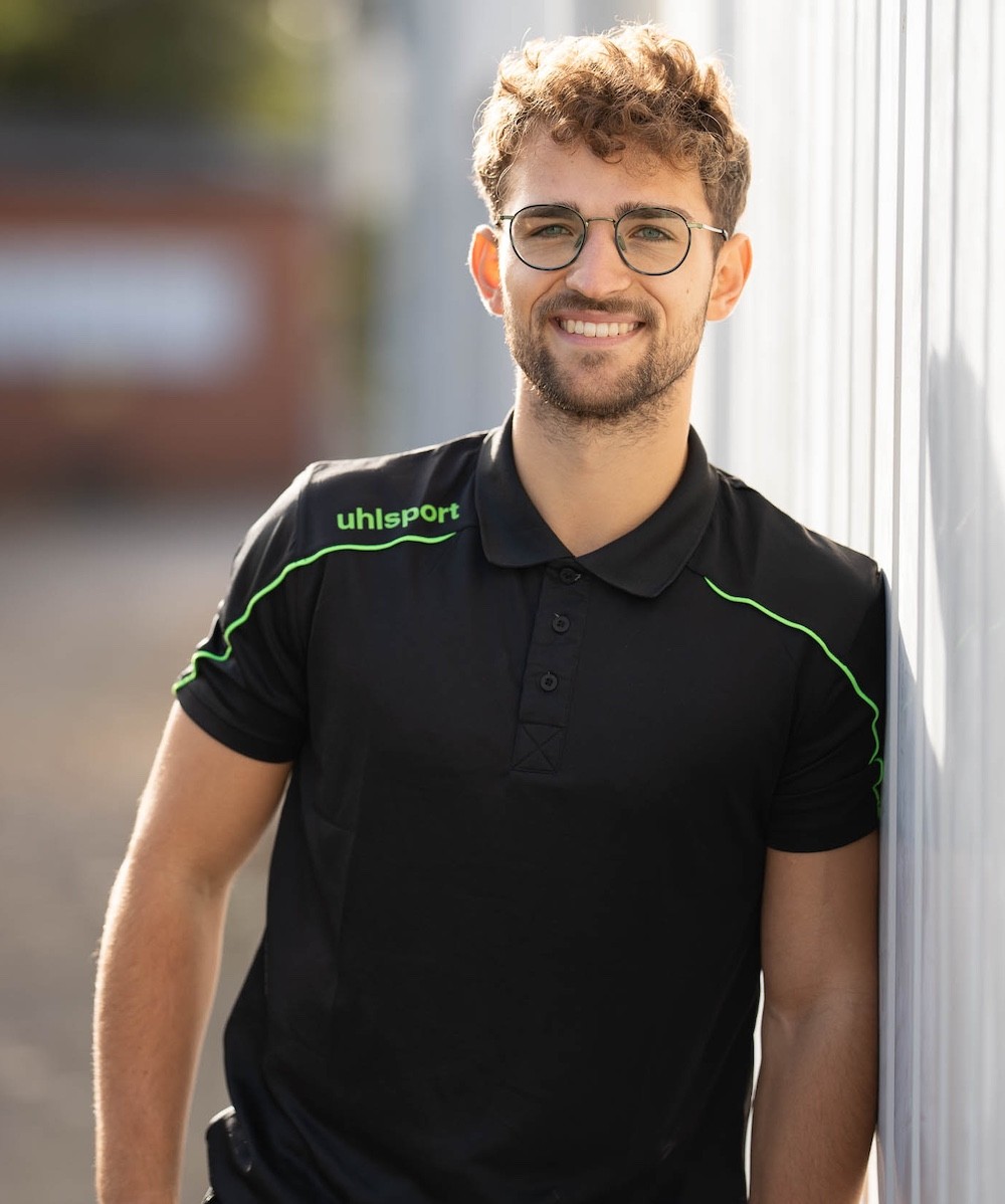Lukas Kazimierski, Personal Trainer aus Hannover, im schwarzen uhlsport Poloshirt vor einer hellen Wand