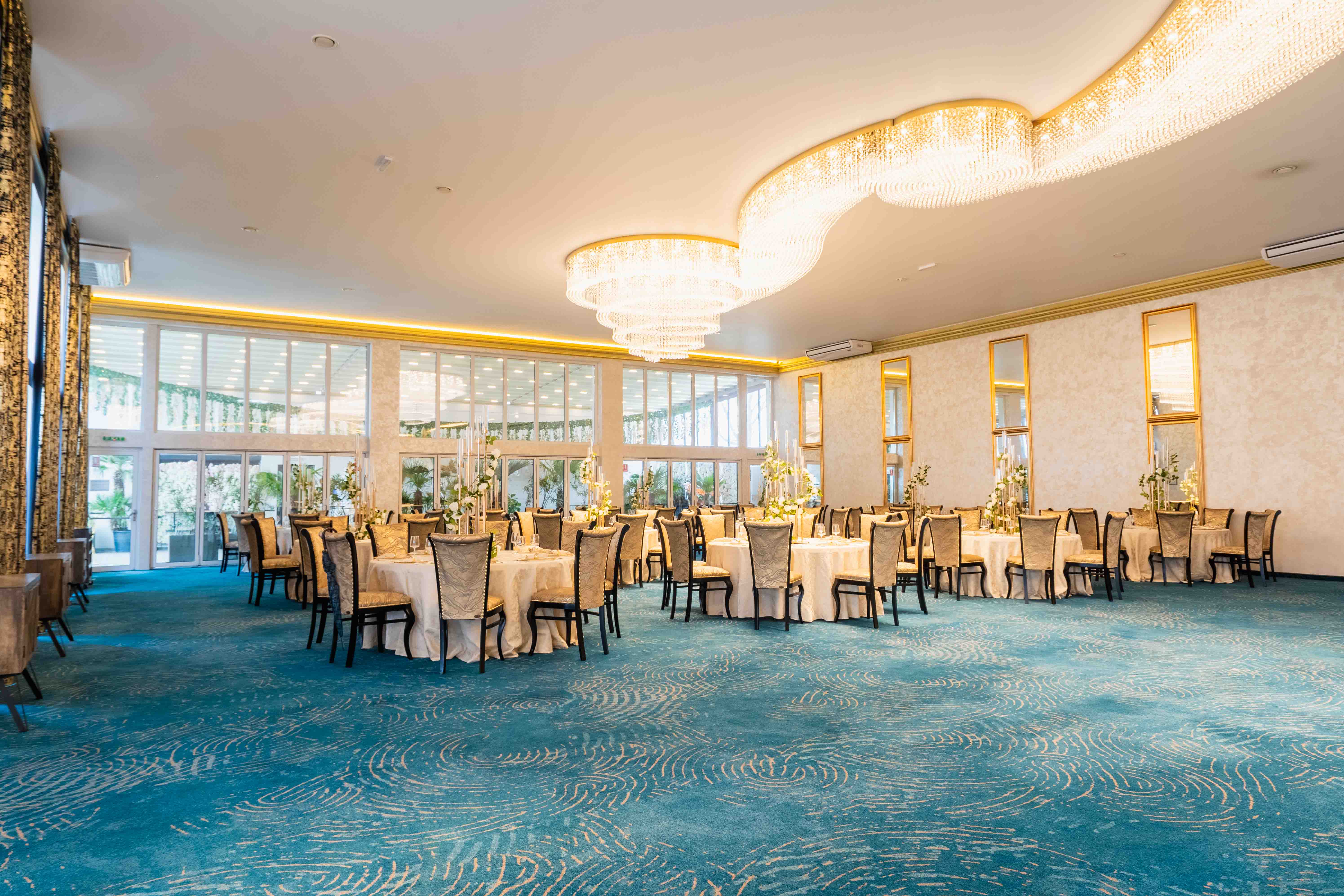 A modern hallway features vibrant teal and orange geometric and leaf patterns, with glossy tiled floors, a large rectangular light fixture, and a small shelf holding a potted plant, all leading to a large, frosted window at the end.