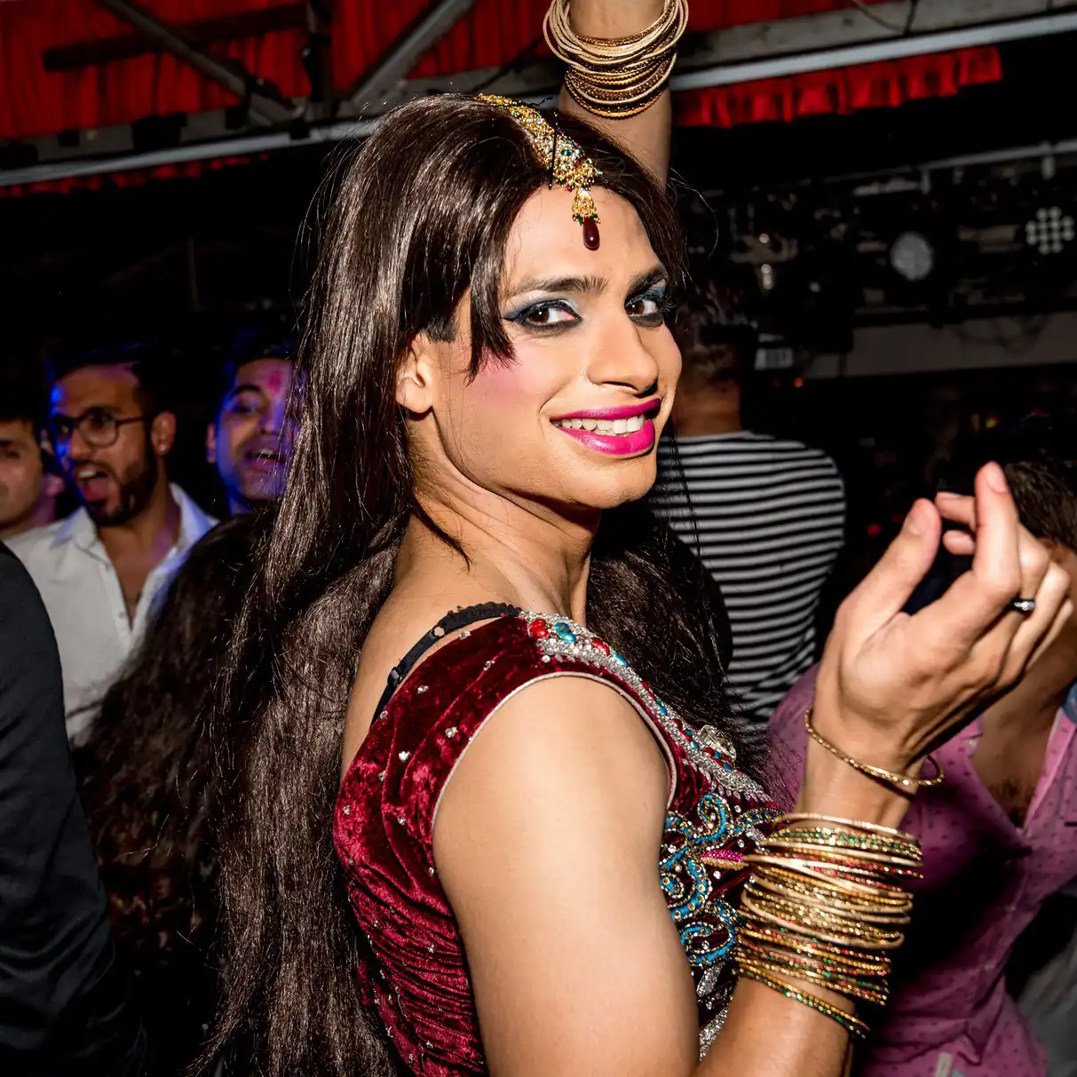 A person smiling exuberantly in a festive setting, wearing colorful makeup, long hair, traditional accessories, and a vibrant outfit. Joyful atmosphere.