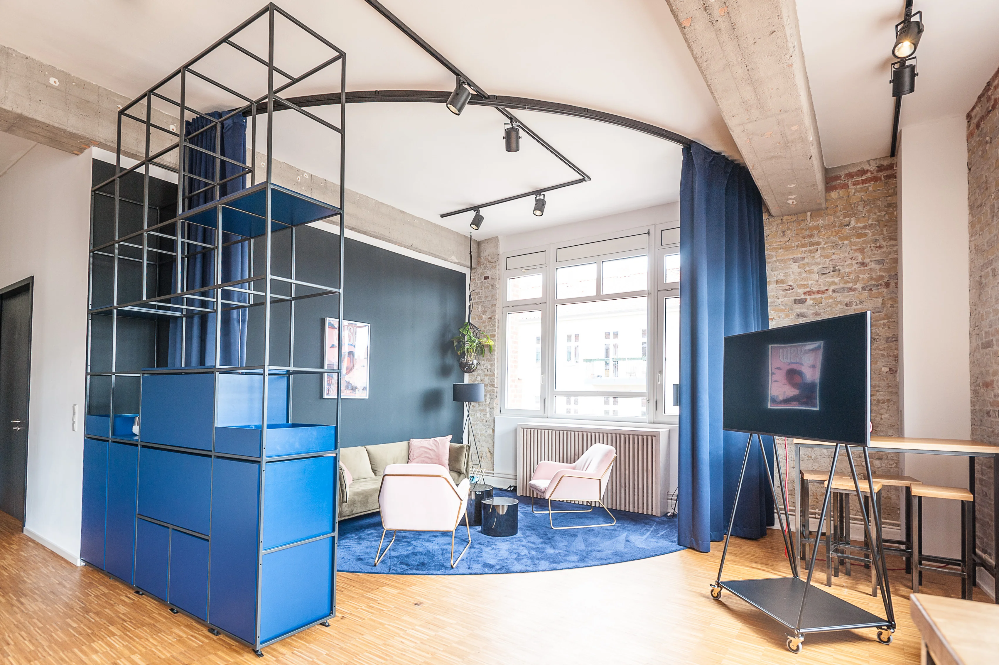 Lounge corner at BASILICOM framed by a curved black track-light arch, with blue modular shelving, dark navy curtains, a beige sofa, pink armchairs, a round blue carpet and a TV on a black easel stand.