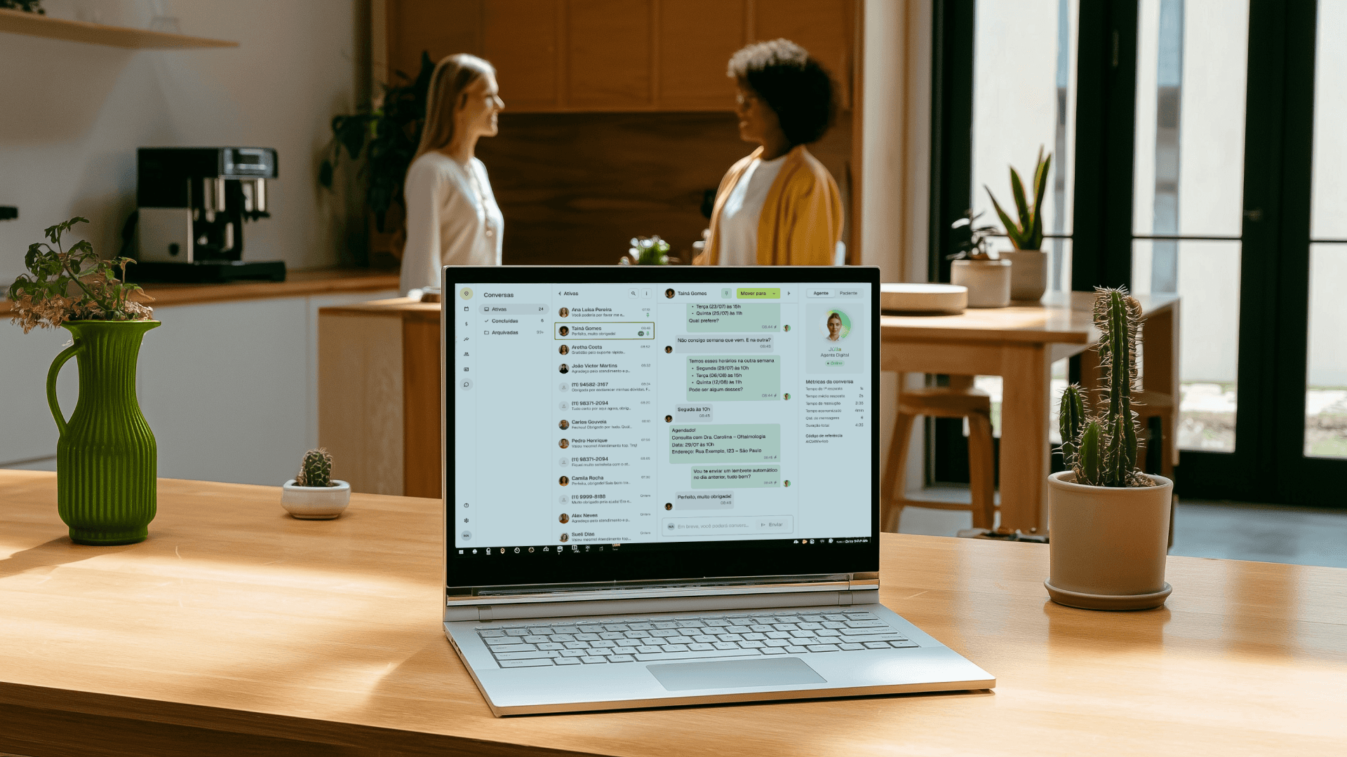 Foto em plano médio de um notebook aberto sobre uma mesa de madeira em uma cozinha moderna e bem iluminada. A tela do notebook exibe a plataforma da Tivita para gestão de clínicas, na aba da Agente Digital Júlia que cuida do WhatsApp da Clínica. No plano de fundo, desfocadas, duas mulheres (uma loira e uma negra) conversam e sorriem. A imagem simboliza a praticidade e a organização do dia a dia de um profissional de saúde que usa a Tivita.
