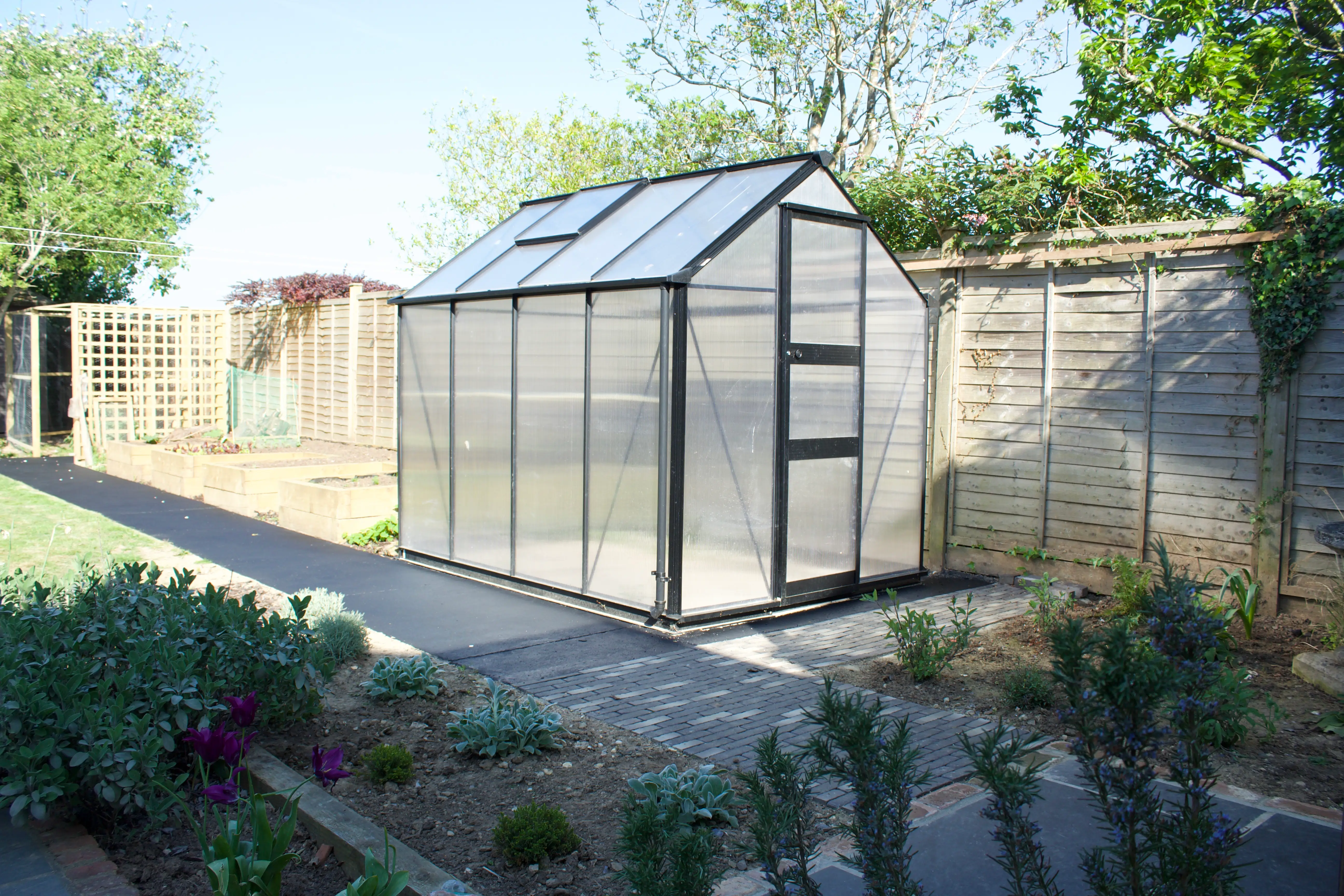 A greenhouse made of glass panels in a garden, surrounded by greenery and a fence. Pathway leads to the entrance.
