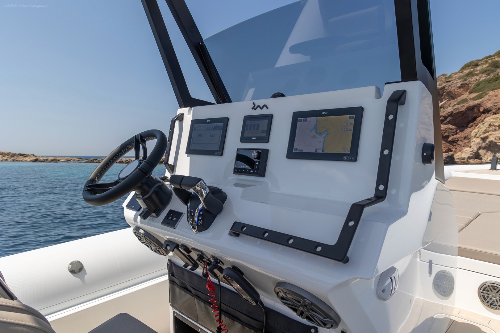 Modern yacht helm station with navigation screens and steering wheel, overlooking calm blue waters and rocky Greek coastline.