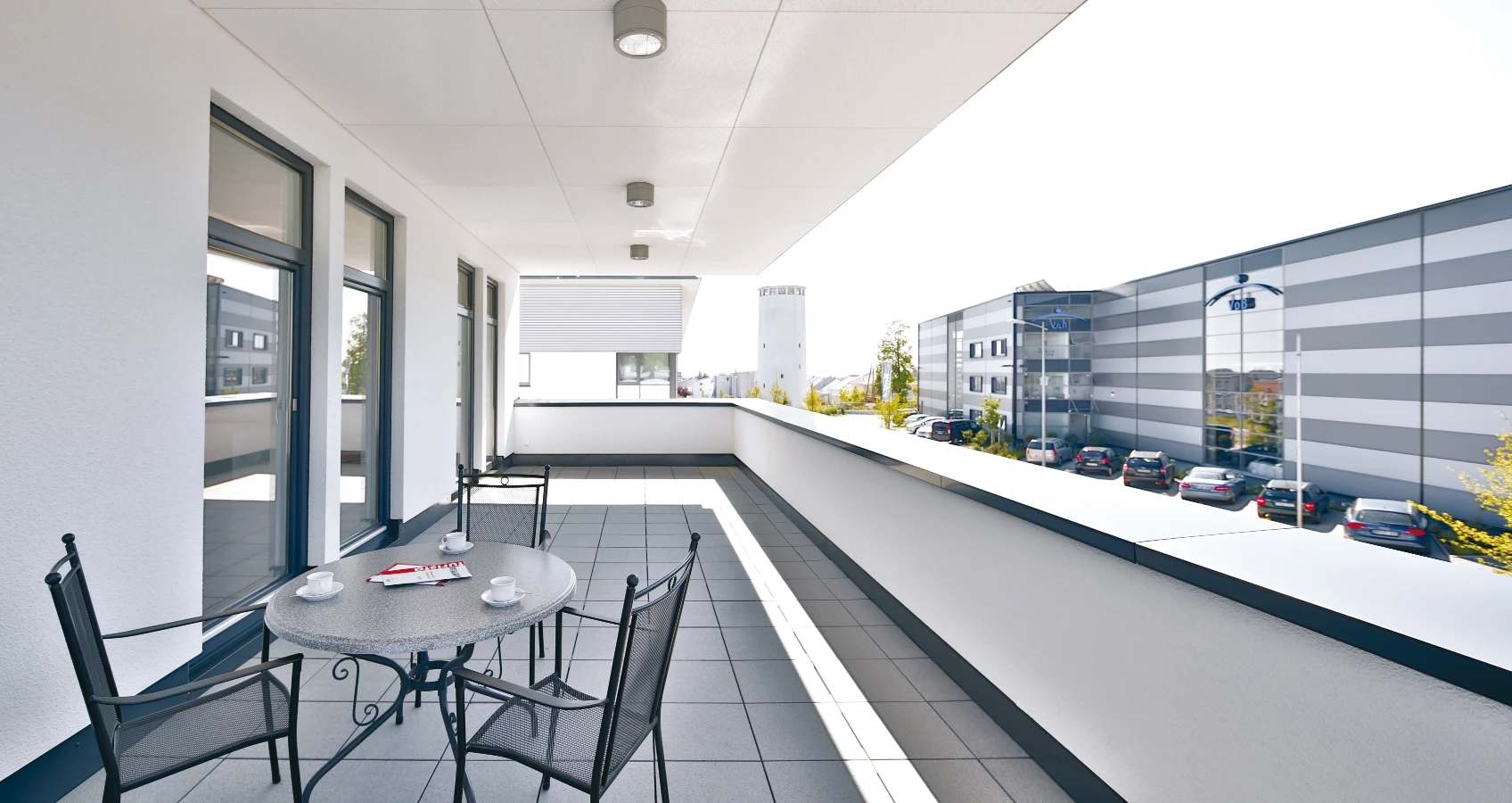 Balkon mit Sitzmöbeln und Kaffeegeschirr vor moderner Industriegebäudearchitektur mit Parkplätzen.