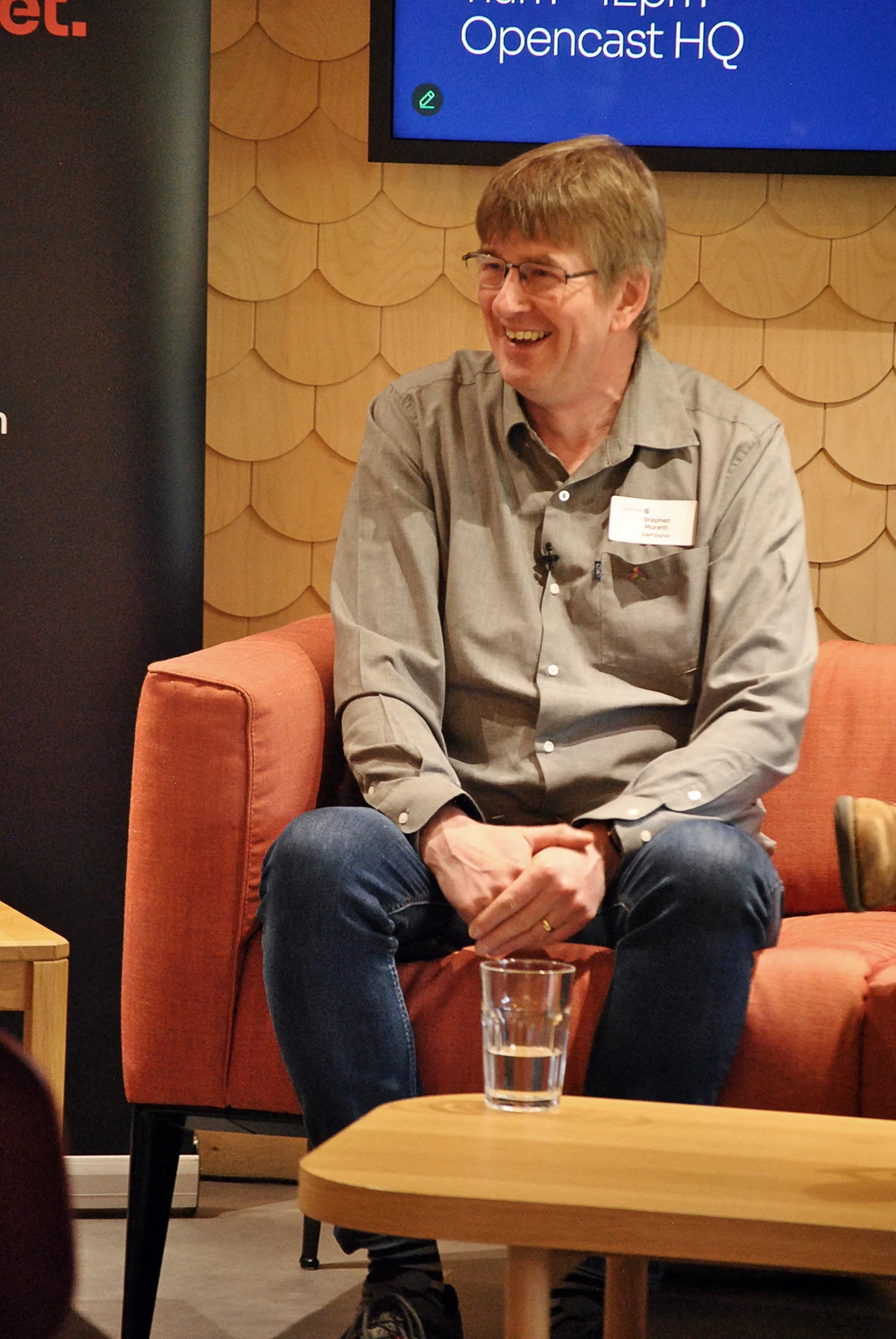 Person seated on an orange armchair with hands clasped, wearing a gray shirt and jeans, with a glass of water on a wooden table in front.