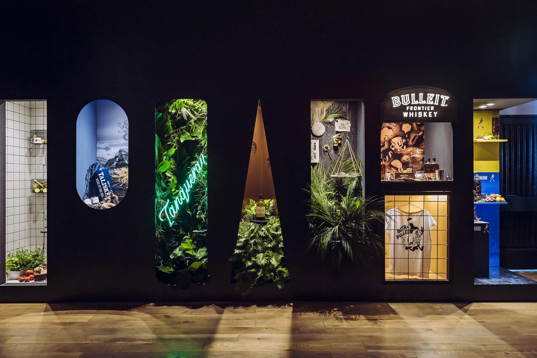 Black display wall at DIAGEO BCB19 with shaped cutout niches showing brand worlds, including a green Tanqueray neon jungle and a Bulleit Whiskey shop window.