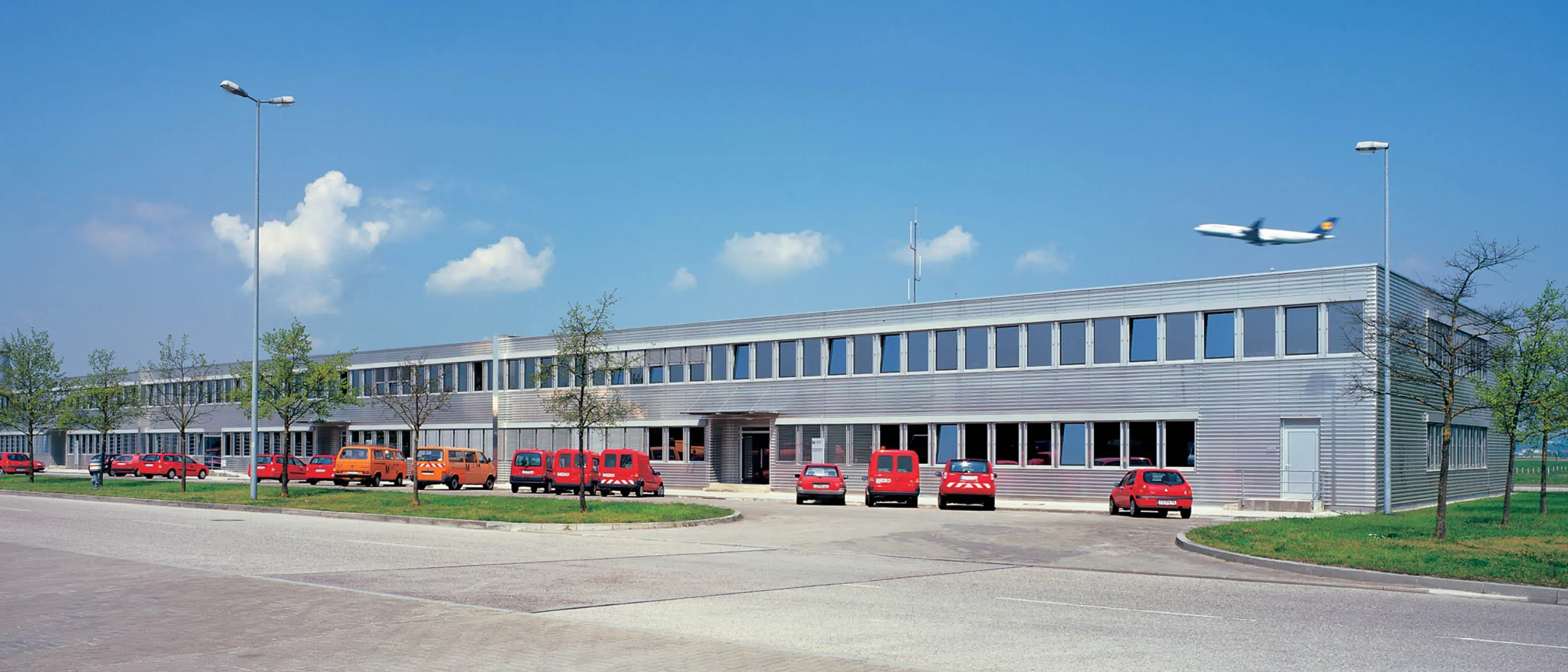 Moderne Industriehalle mit geparkten roten und orangen Lieferwagen. Ein Flugzeug fliegt am Himmel vorbei.