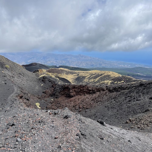 Paisagem vulcânica com terreno rochoso e crateras sob um céu parcialmente nublado. Montanhas e vales distantes são visíveis.