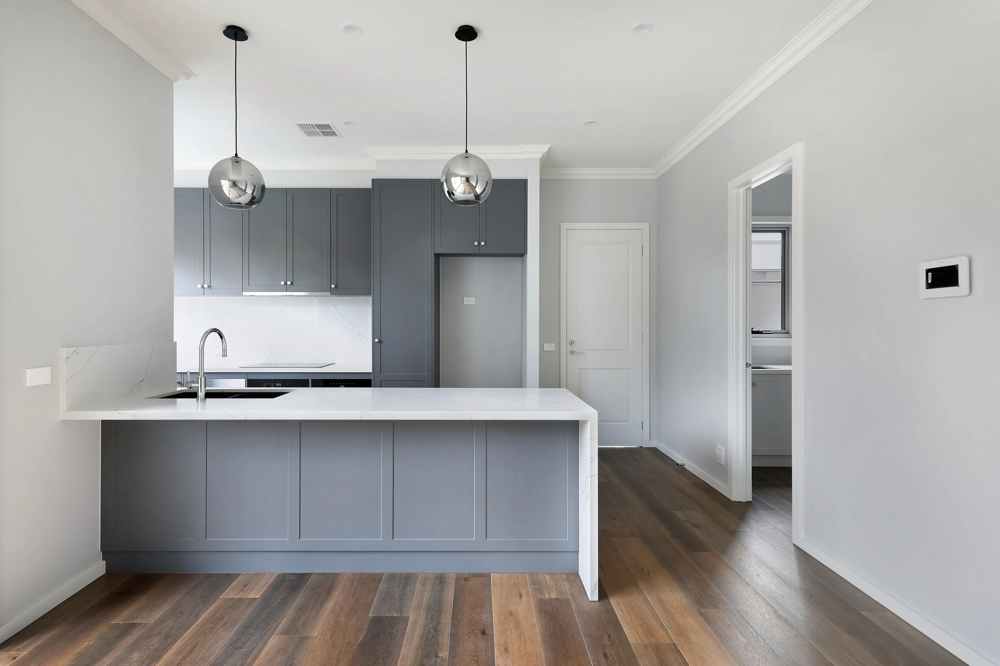 Grey shaker kitchen with a white waterfall stone island, marble splashback, chrome sphere pendants and oak floors, with a hallway leading to the butler's pantry