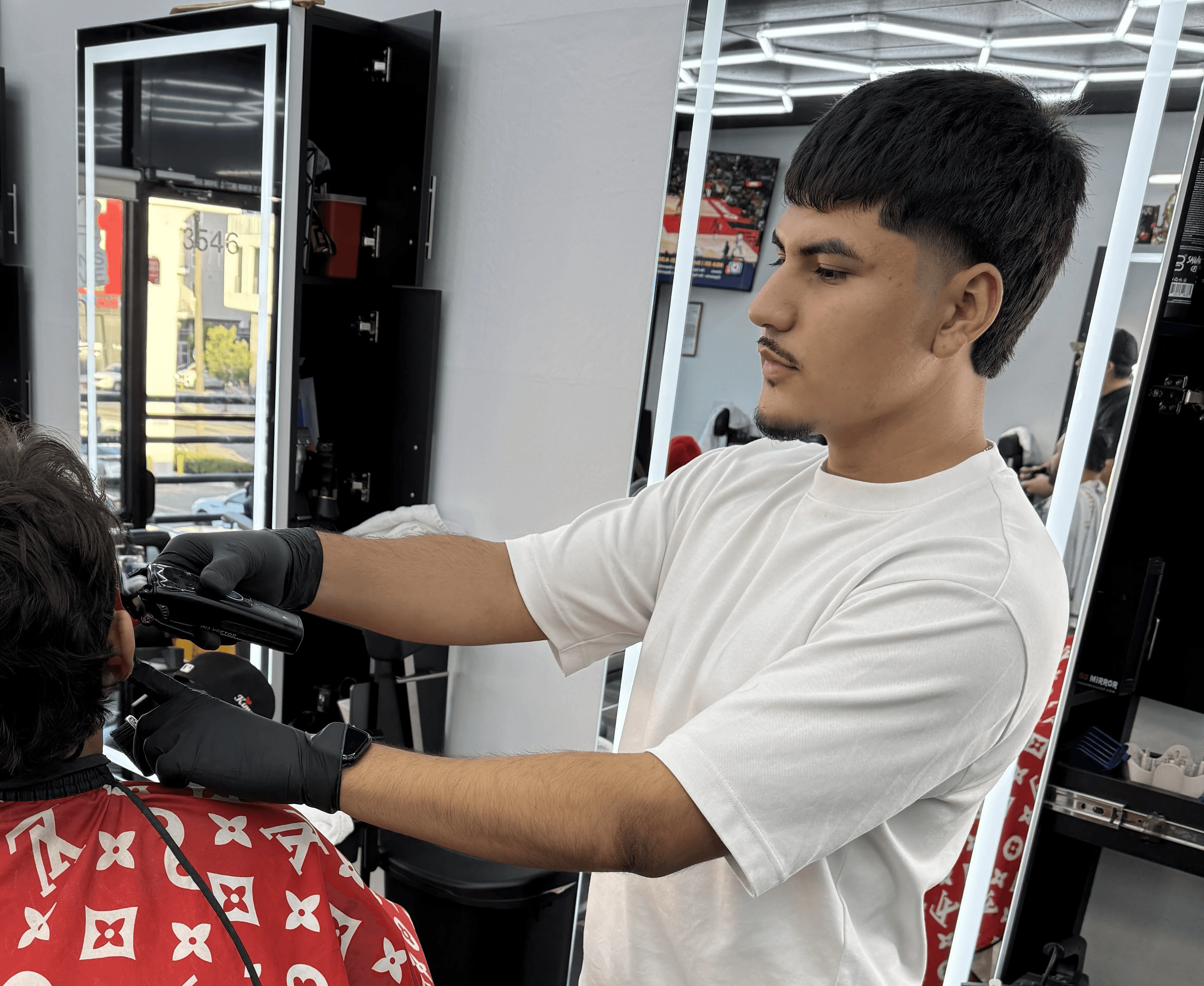 Barber standing with arms crossed in a modern barbershop, wearing a black cap, black T-shirt, and white pants, with hexagon ceiling lights in the background.