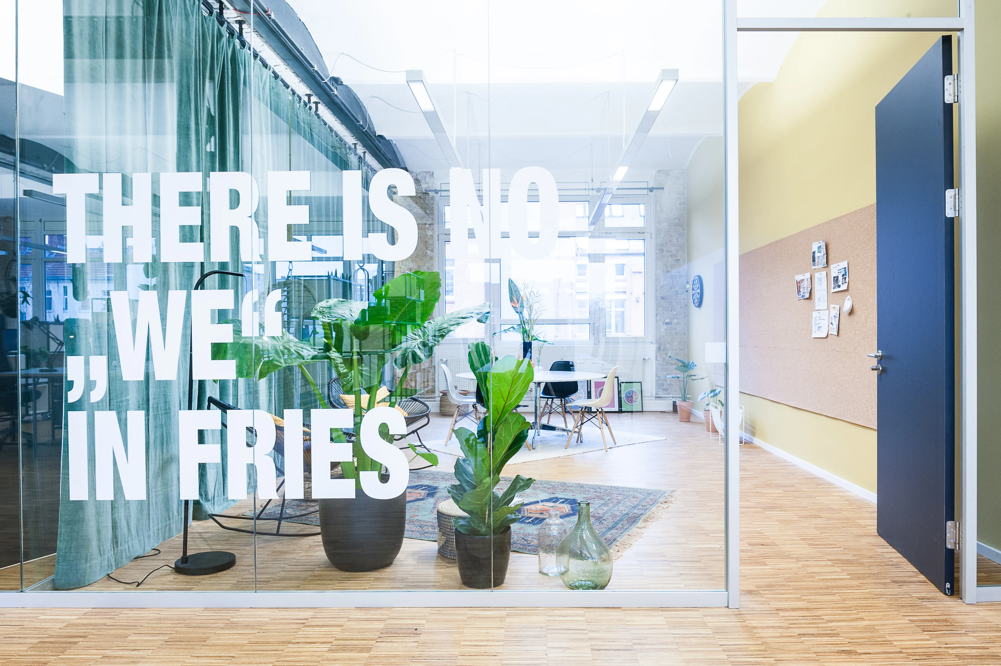 Glass entrance wall at INTERMATE x TRUEMATES with the white slogan "THERE IS NO WE IN FRIES", a green velvet curtain, potted plants and a meeting corner with cork pinboard behind a blue door.