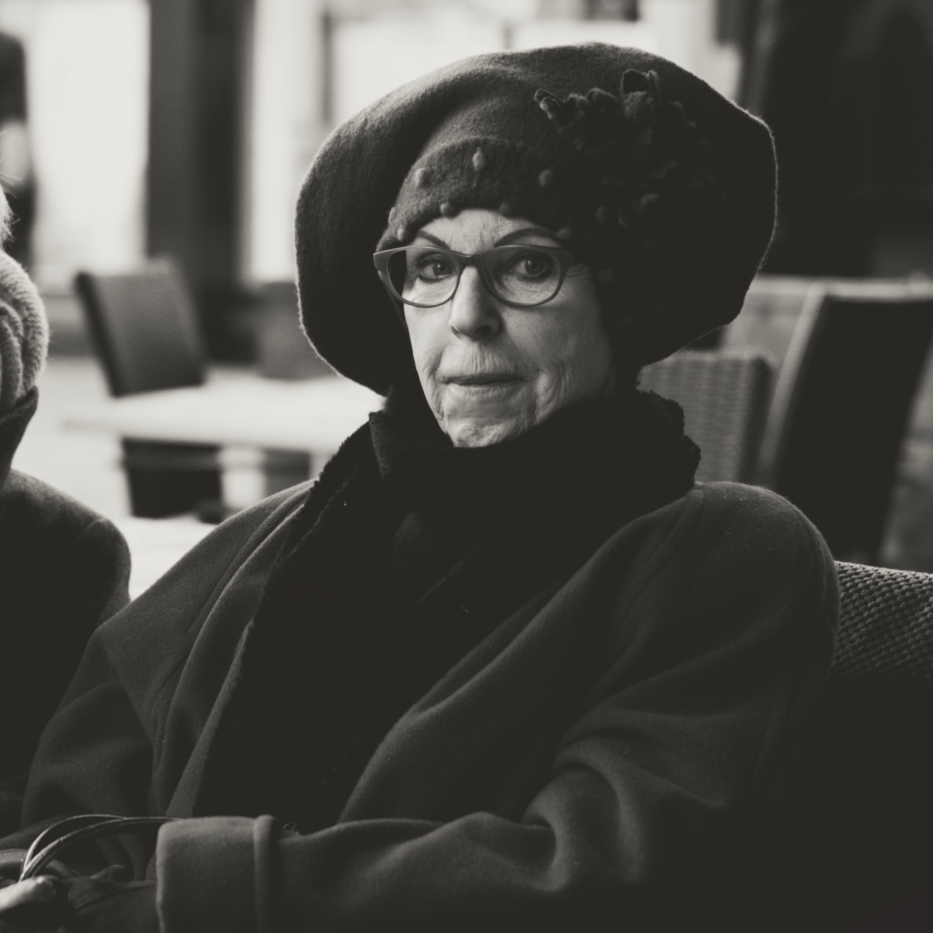 Black-and-white café portrait of an older person in a textured hat and coat — ideal for street photography, portraiture, vintage style, and lifestyle blog content about urban cafés and timeless portraits.