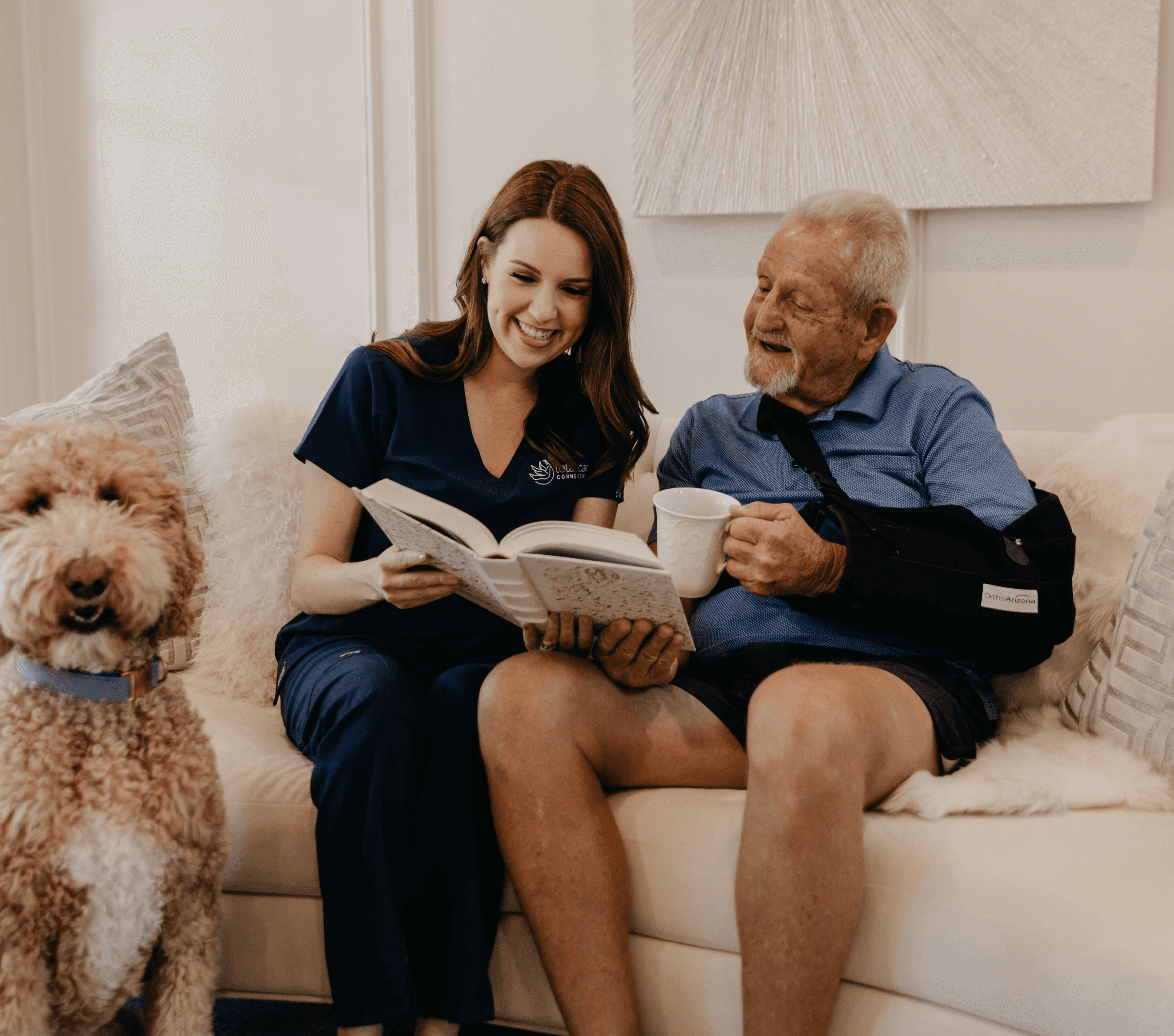 A woman and man sit on a couch with a dog beside them, looking at a book.