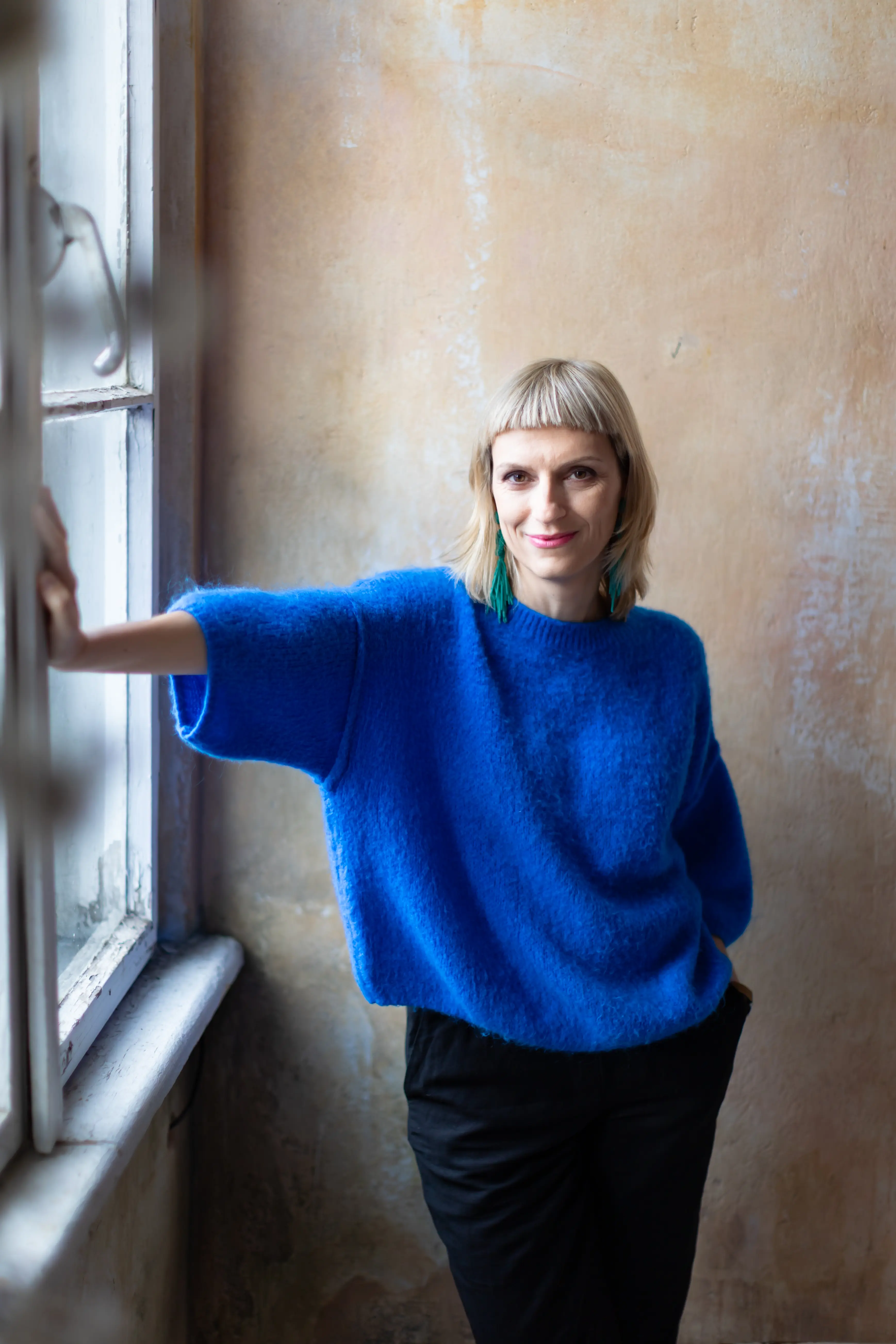 Osoba o krótkich blond włosach ubrana w jasnoniebieski sweter i czarne spodnie stoi w pomieszczeniu przy oknie, uśmiechając się z jedną ręką opartą na parapecie na tle beżowej ściany.
