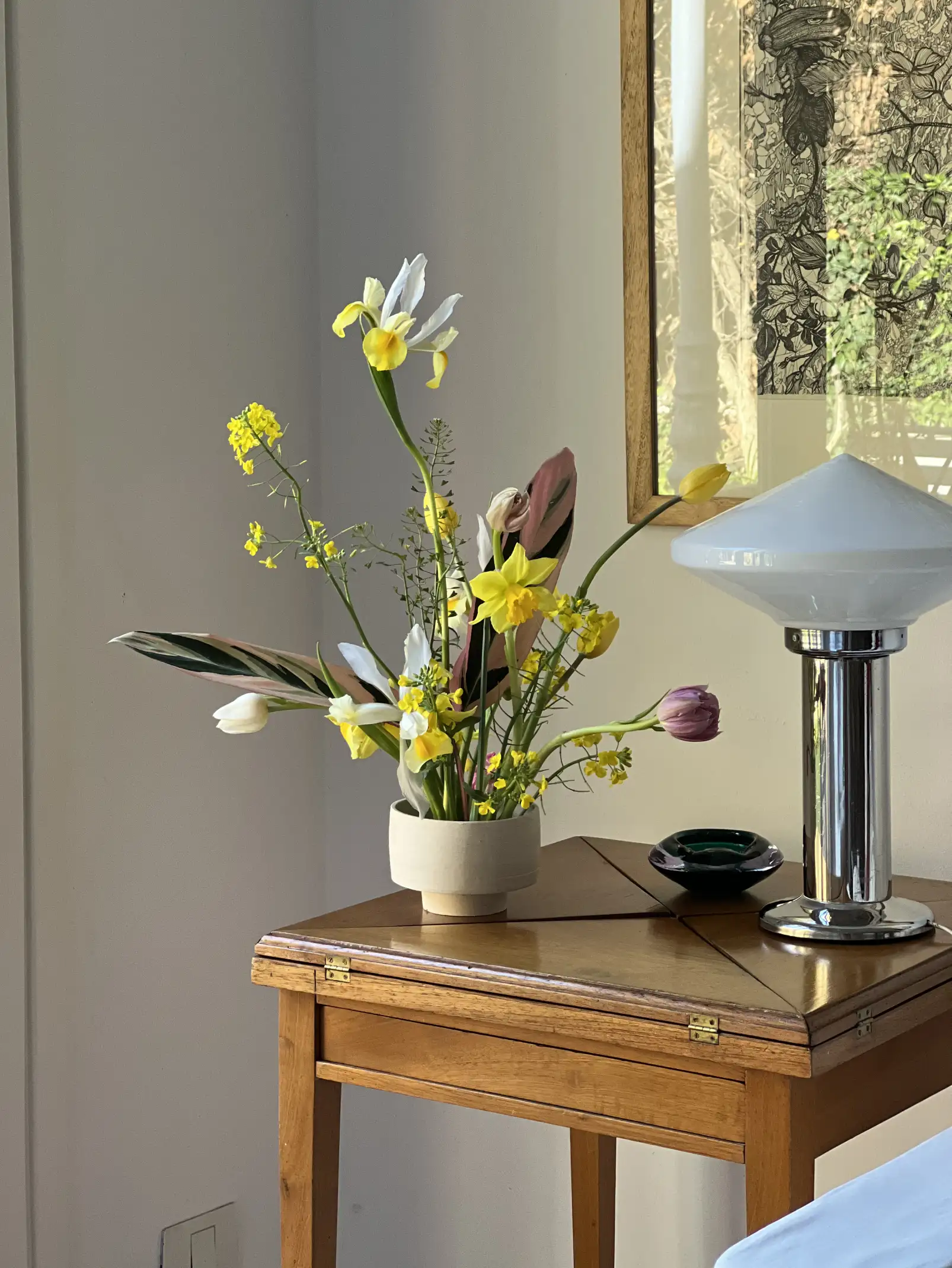 Sala luminosa con sillón blanco, cojines verdes y un aparador de madera decorado con un florero de ramas con flores rosas, cuadros en la pared y piso de madera natural