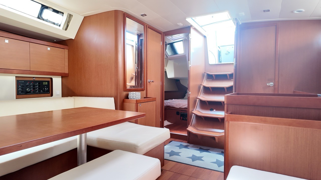 Interior view of the Beneteau Oceanis 45's saloon, showing a mahogany dining table with cream cushioned bench seating, a navigation panel, a mirrored door, and a companionway staircase leading to the forward cabin over a star-patterned rug.