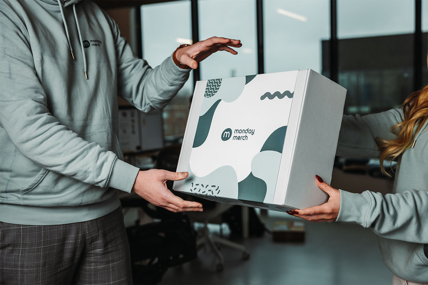 Twee mensen in een kantoor, die een doos vasthouden met het merk "Monday Merch"