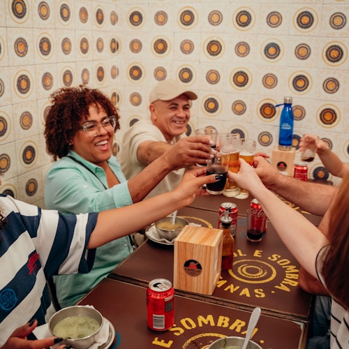 四个人在餐桌上举杯庆祝，脸上挂着微笑。桌上有罐子和碗。墙壁上覆盖着有图案的瓷砖。