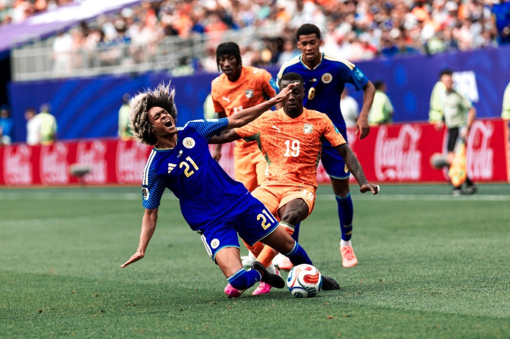 A Curaçao player falls after a challenge while Ivory Coast's number 19 controls the ball near the Coca-Cola boards