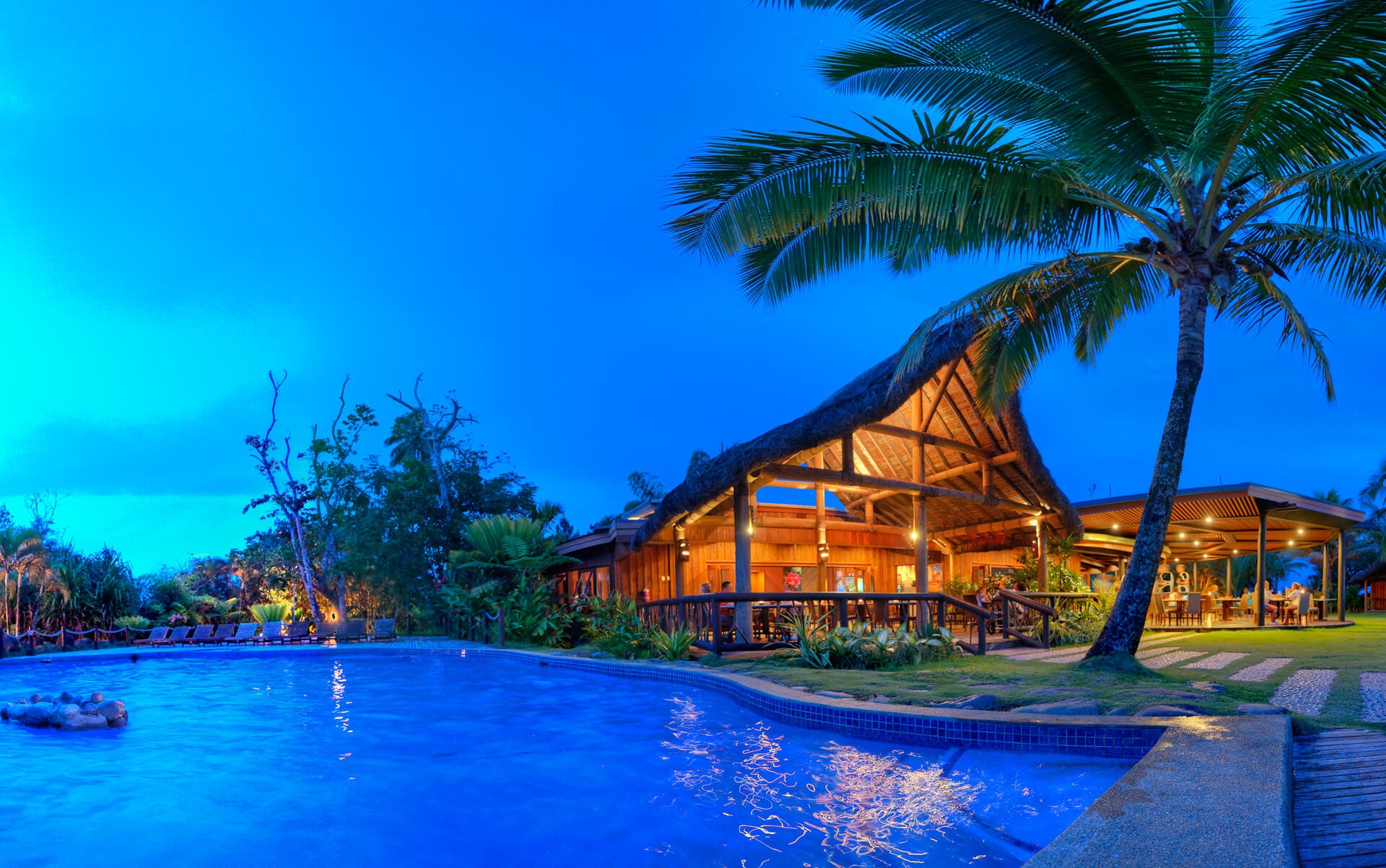 Uprising Beach Resort dining pavilion at dusk with a giant palm tree in front and the pool in the foreground