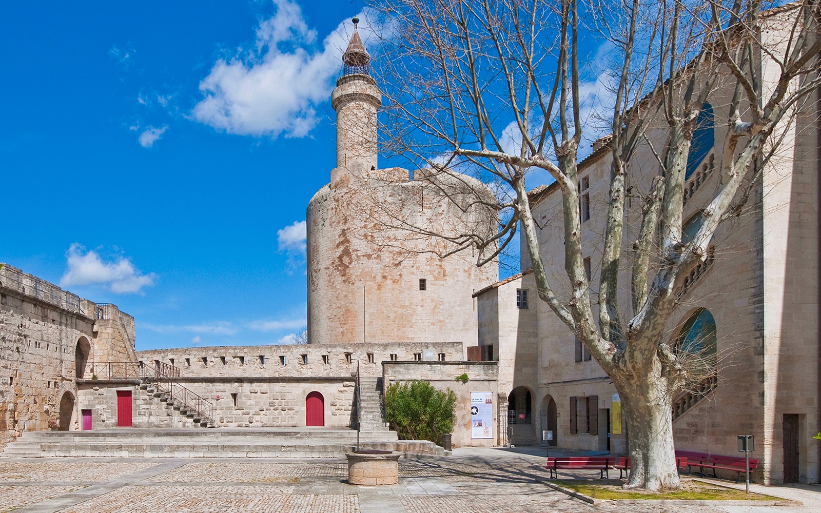 Aigues-Mortes tower and ramparts with stone courtyard and tree under blue sky.