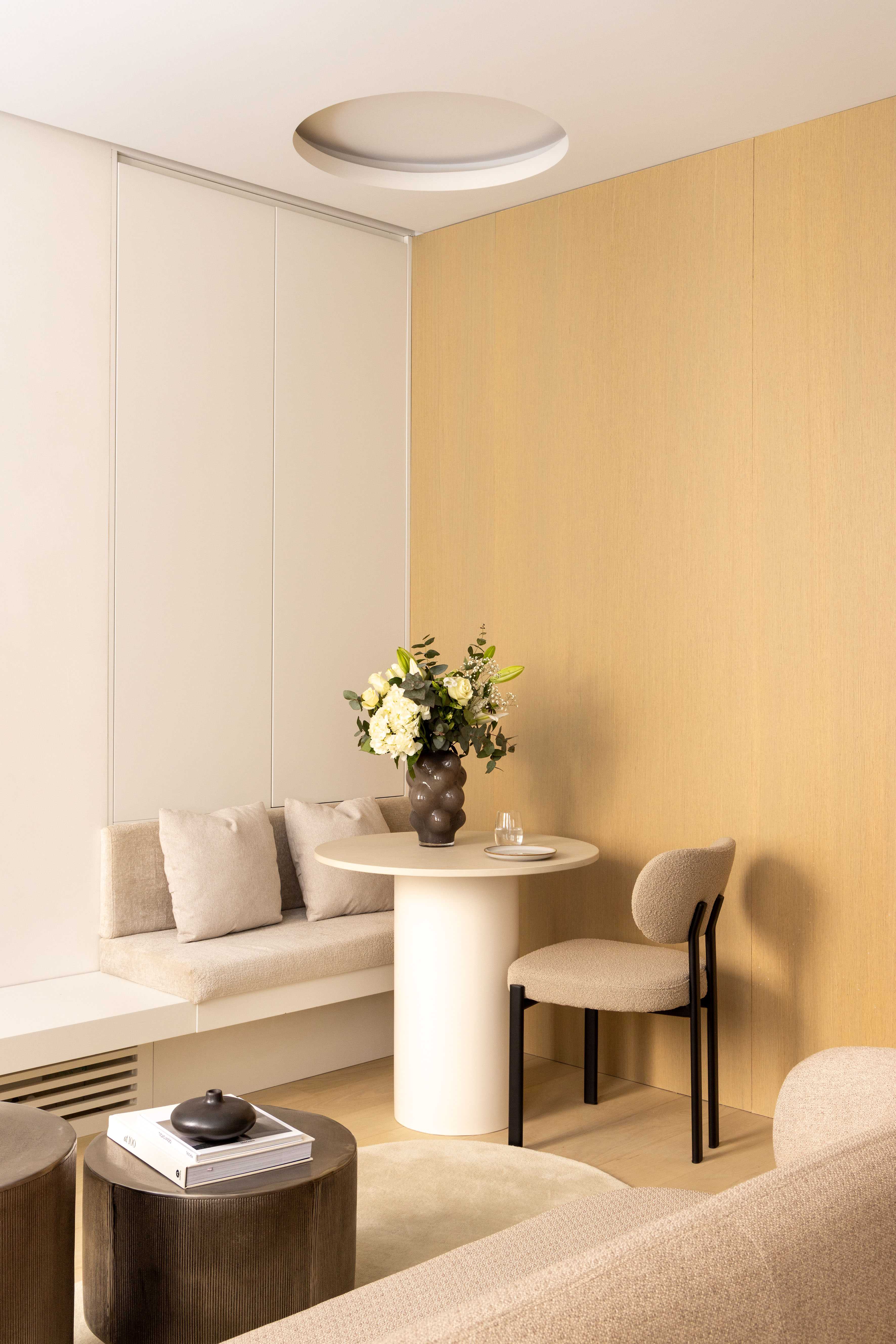 Coin repas avec banquette beige, table ronde blanche, chaise bouclette et bouquet de fleurs blanches
